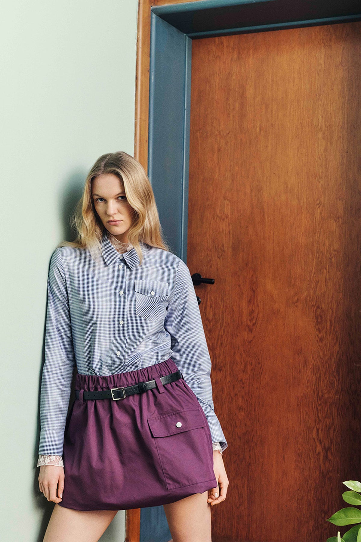 Model beside a wooden door wearing a blue-and-white checked shirt with lace trim and a belted plum utility skirt with a flap pocket