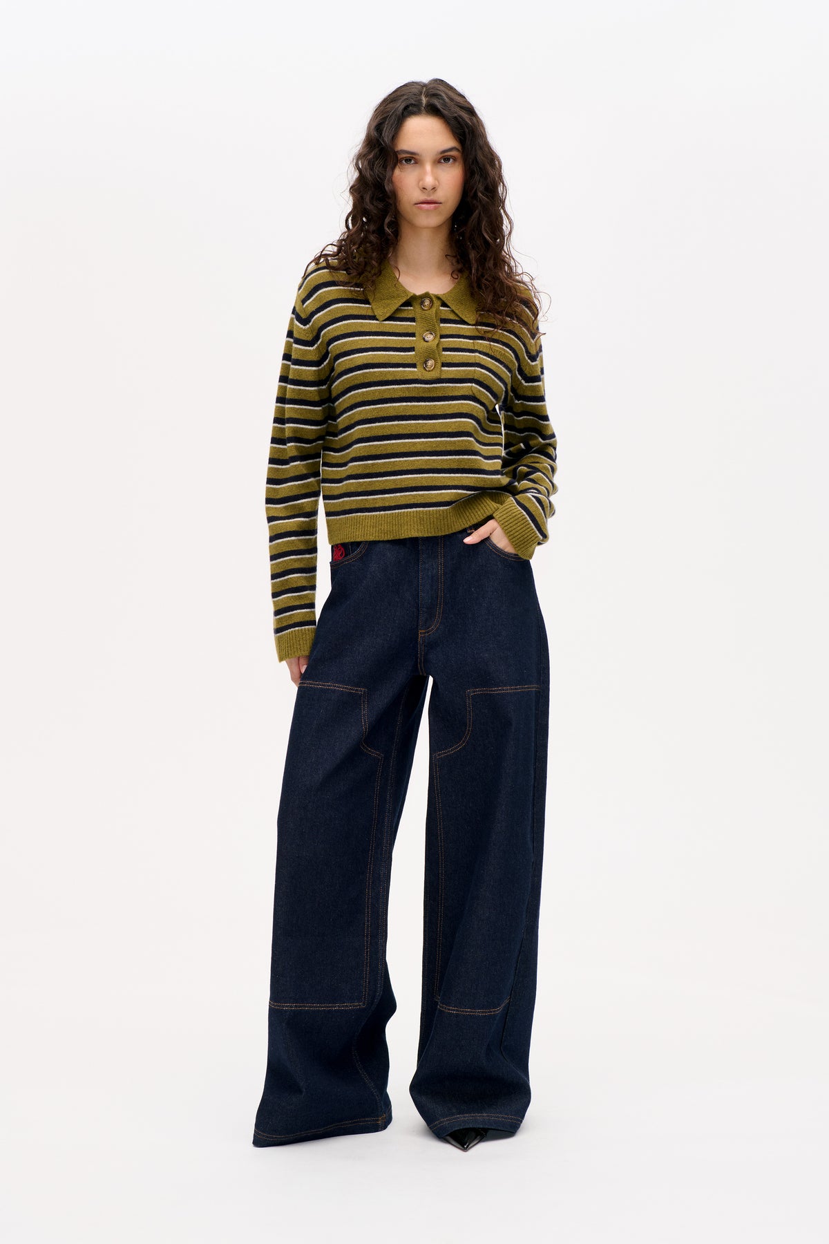 Model trägt einen olivgrünen, gestreiften, langärmligen Strickpullover mit spitzem Kragen und Knopfleiste, kombiniert mit dunklen, weiten Jeans.