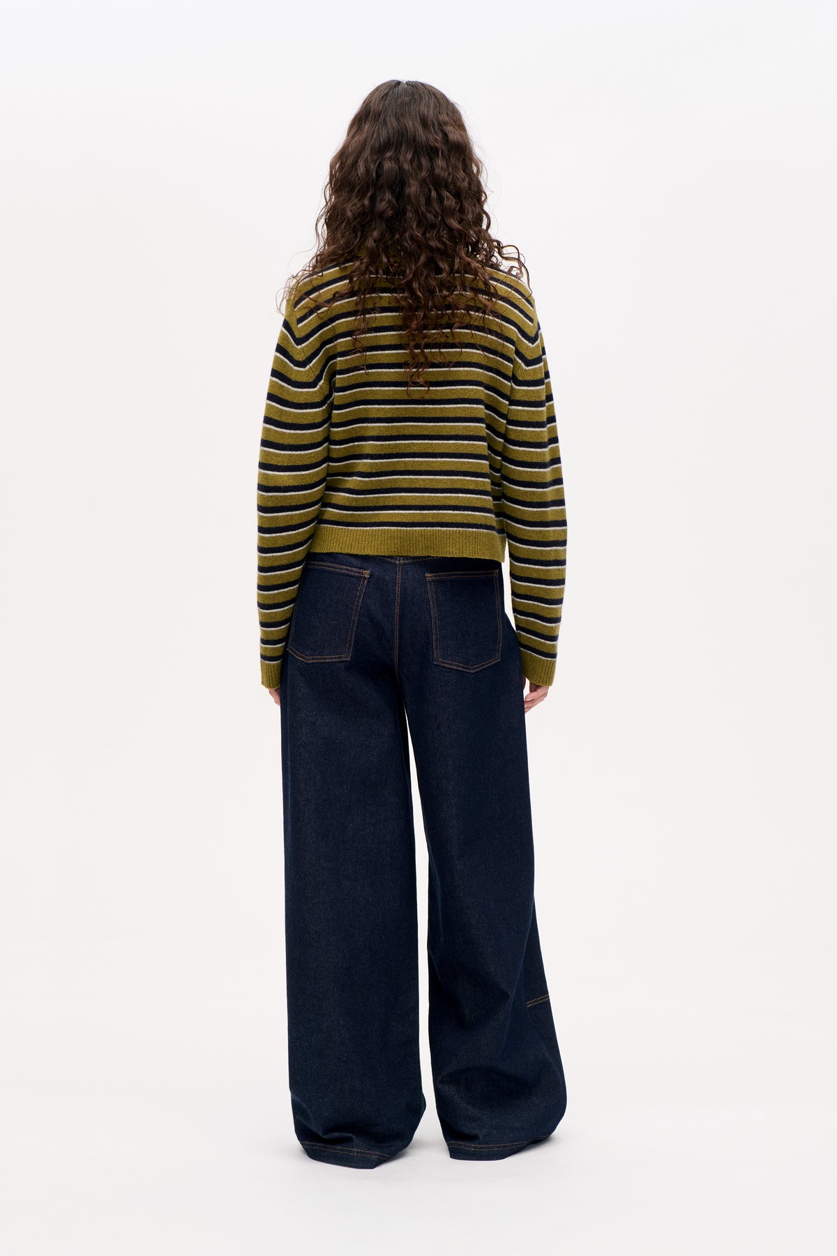 Back view of a model wearing an olive green, navy and white striped sweater with dark wide-leg jeans.