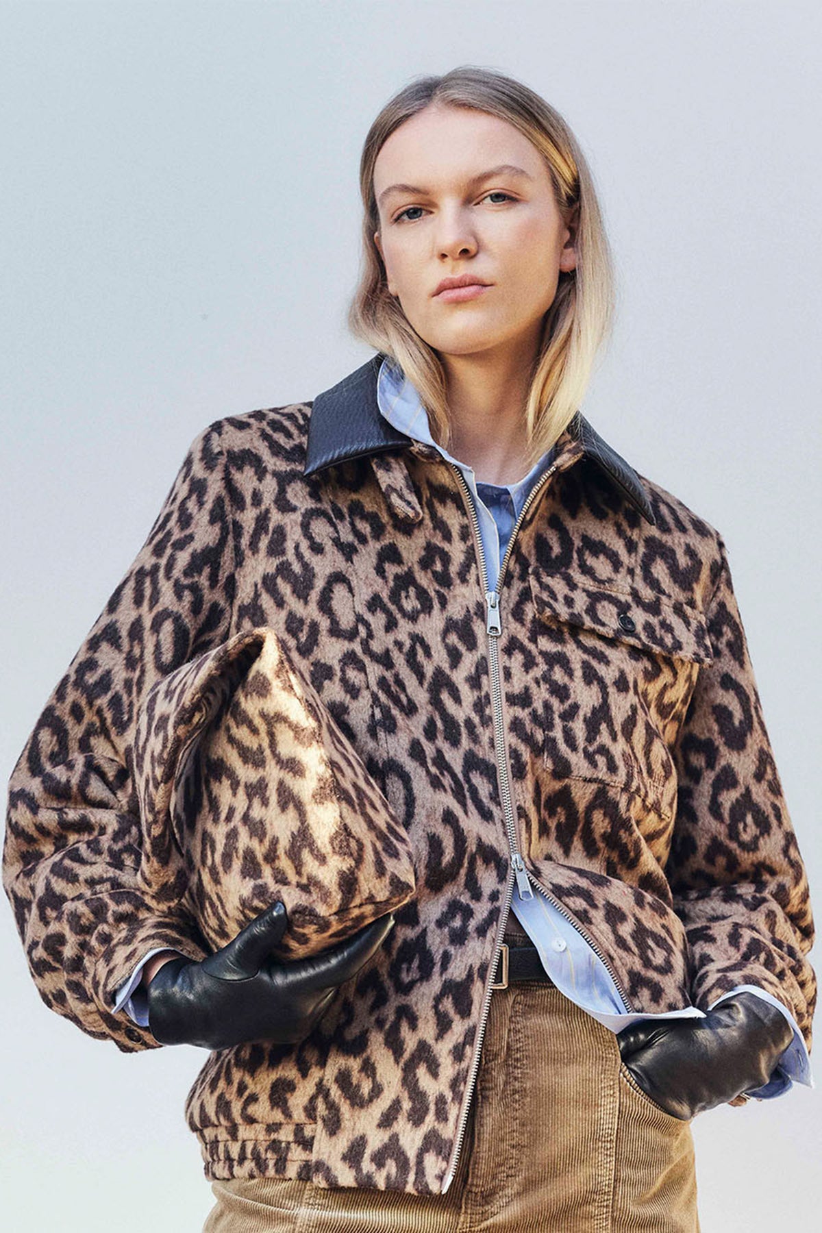 Fashion campaign portrait featuring a brown and black leopard-print zip-front jacket layered over a blue shirt and brown ribbed top.
