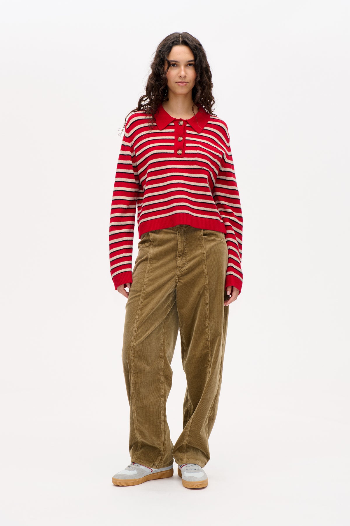 Front view of a model wearing a red knit sweater with cream and navy stripes, a pointed collar and button placket, paired with brown corduroy trousers.