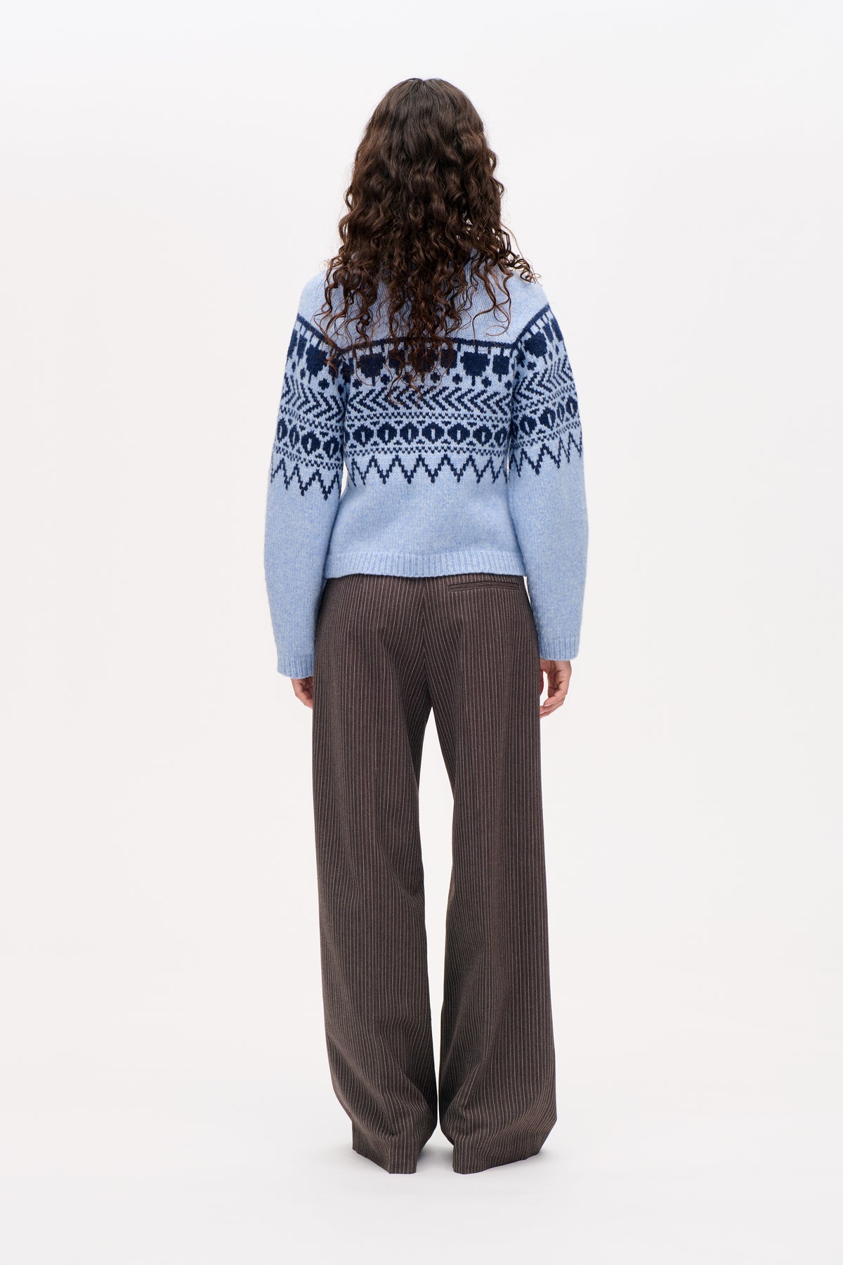 Vue de dos d'un pull bleu clair à motifs géométriques Fair Isle bleu marine foncé, porté avec un pantalon large marron foncé à fines rayures.