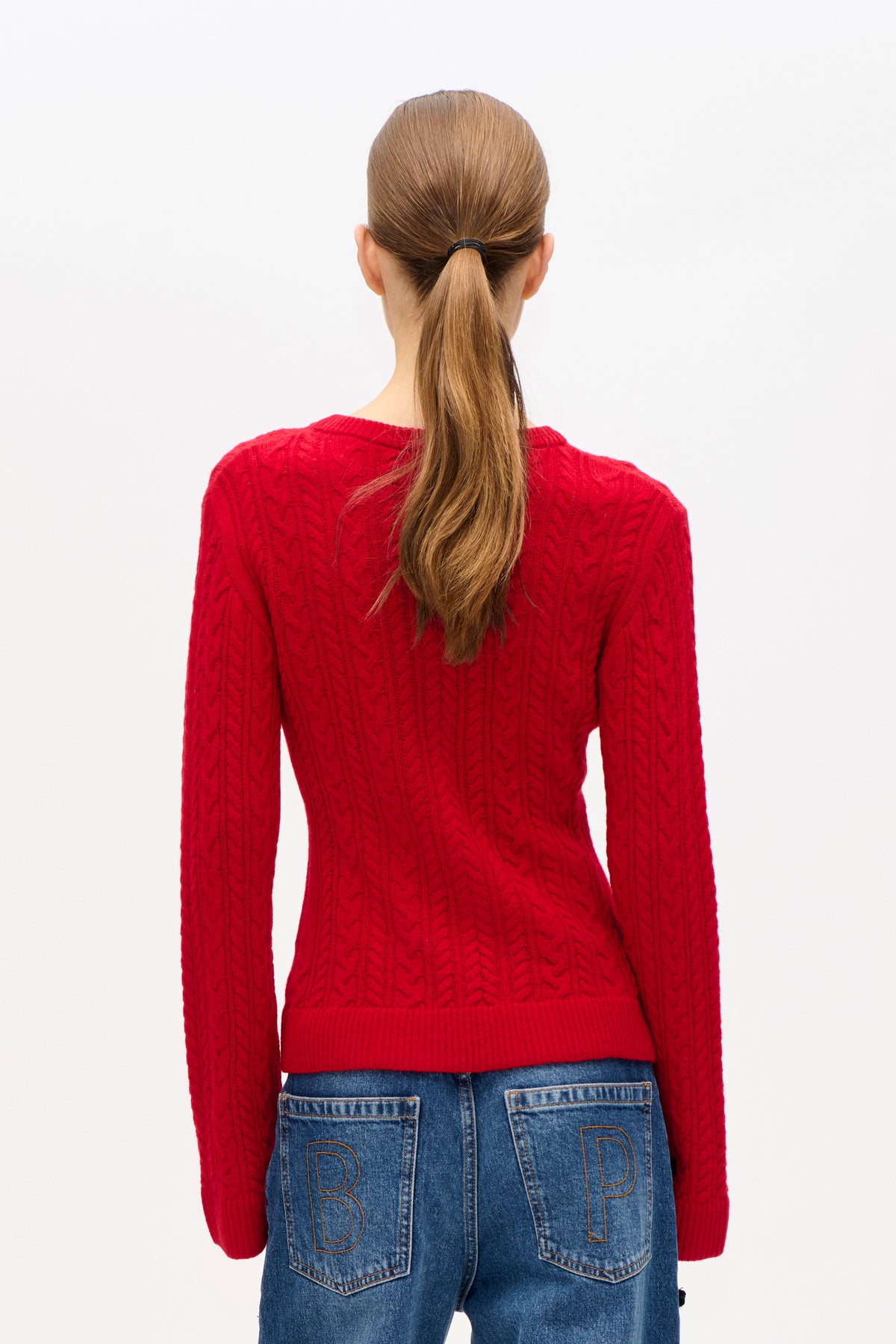 Back view of a model wearing a bright red cable-knit sweater and blue jeans with outlined letters on the back pockets.