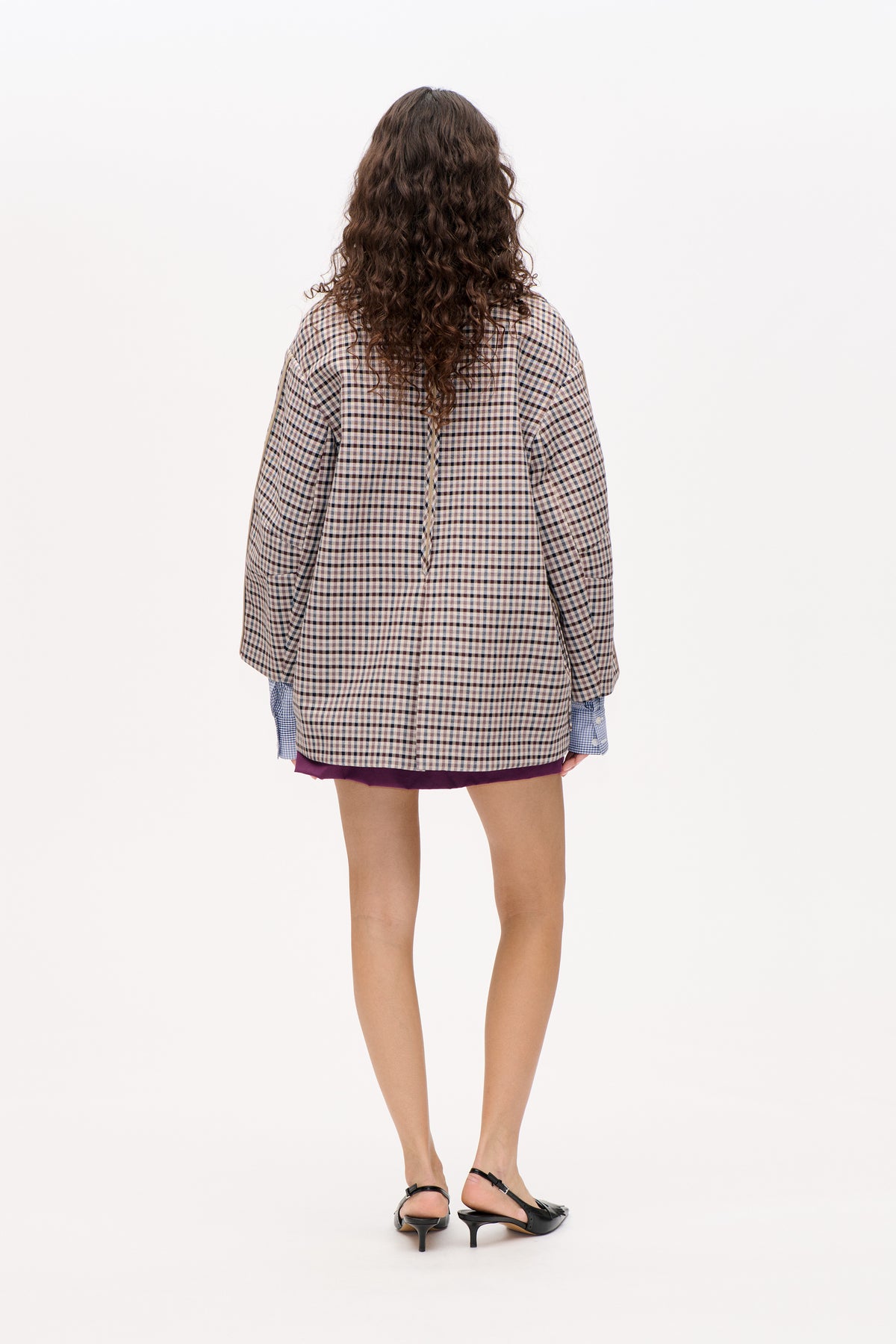 Back view of an oversized beige, brown and navy checked jacket, layered over a purple skirt and blue checked shirt.
