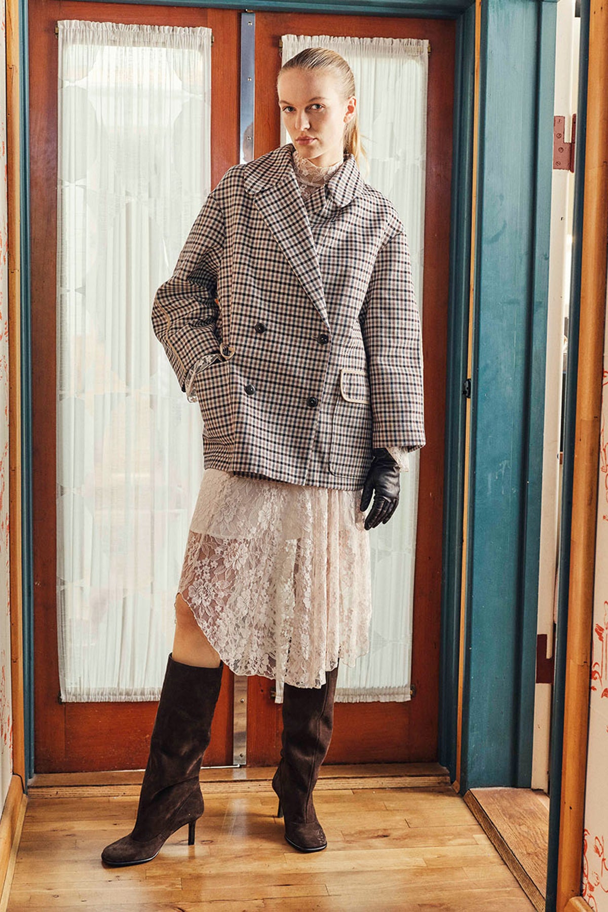 Model in a black-and-cream checked double-breasted jacket over a pale lace dress, with dark gloves and knee-high boots, framed in a doorway.