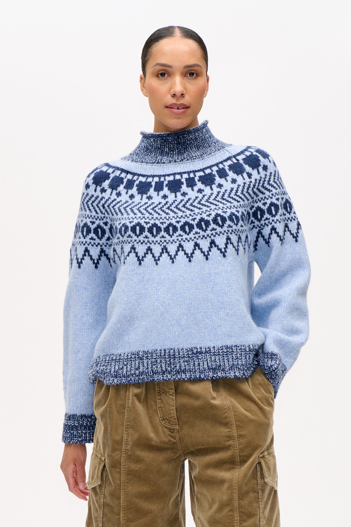 Vooraanzicht van een model dat een lichtblauwe gebreide trui draagt met een hoge geribde kraag en een marineblauw geometrisch patroon over de schouders.