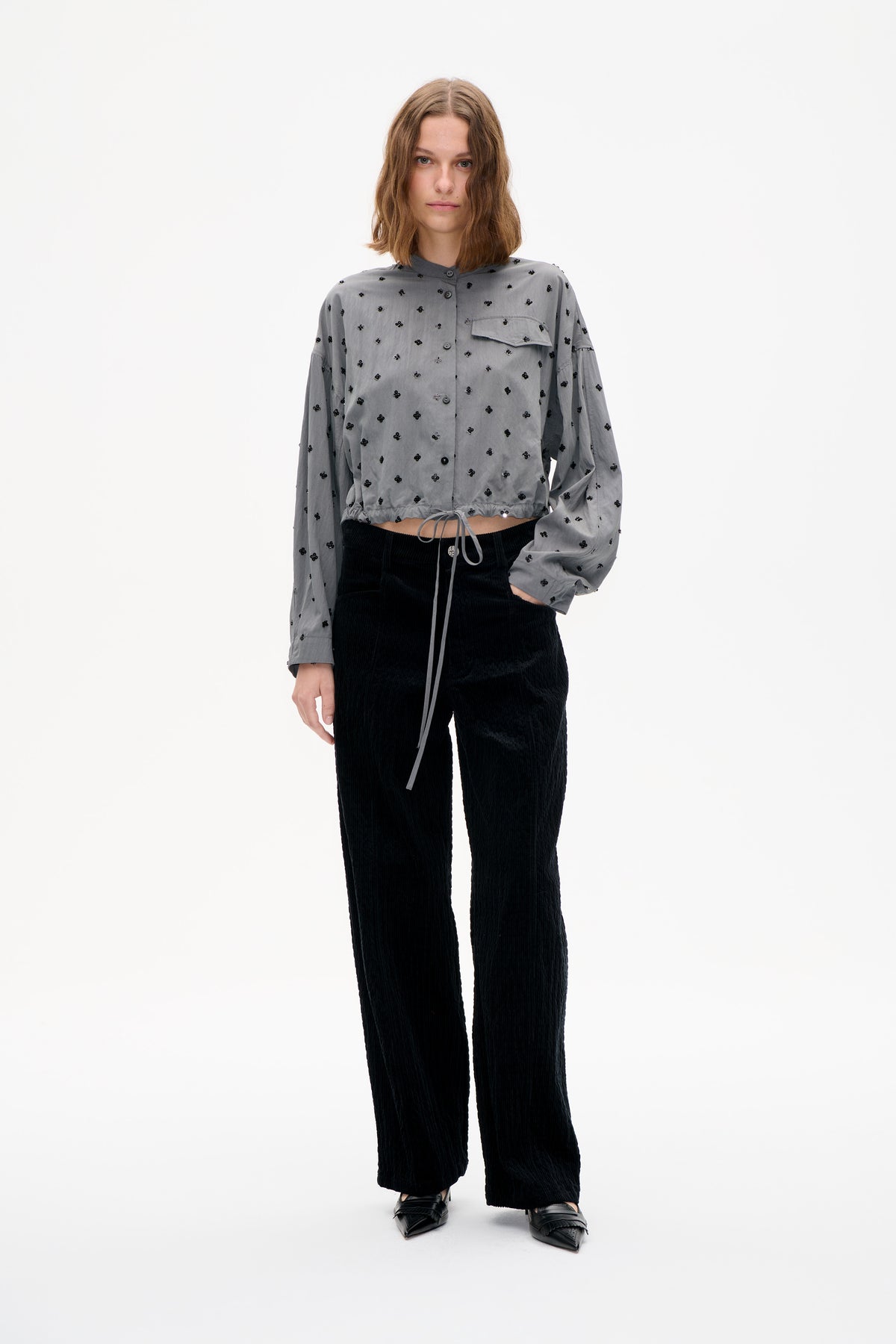 Frontalansicht eines Models in einem kurzen grauen Hemd mit Knopfleiste, schwarzen floralen Applikationen, Brusttasche und Kordelzugsaum, kombiniert mit schwarzen Hosen.