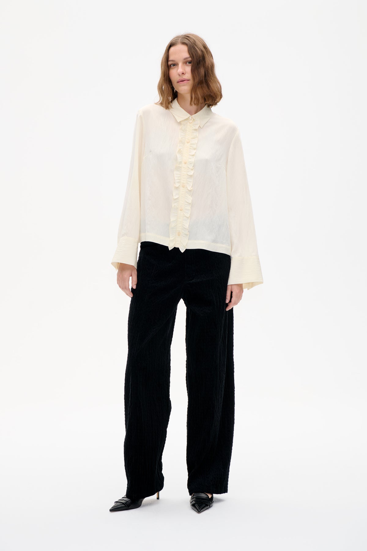 Model wearing an ivory long-sleeved blouse with a pointed collar, ruffled button placket and black textured wide-leg trousers