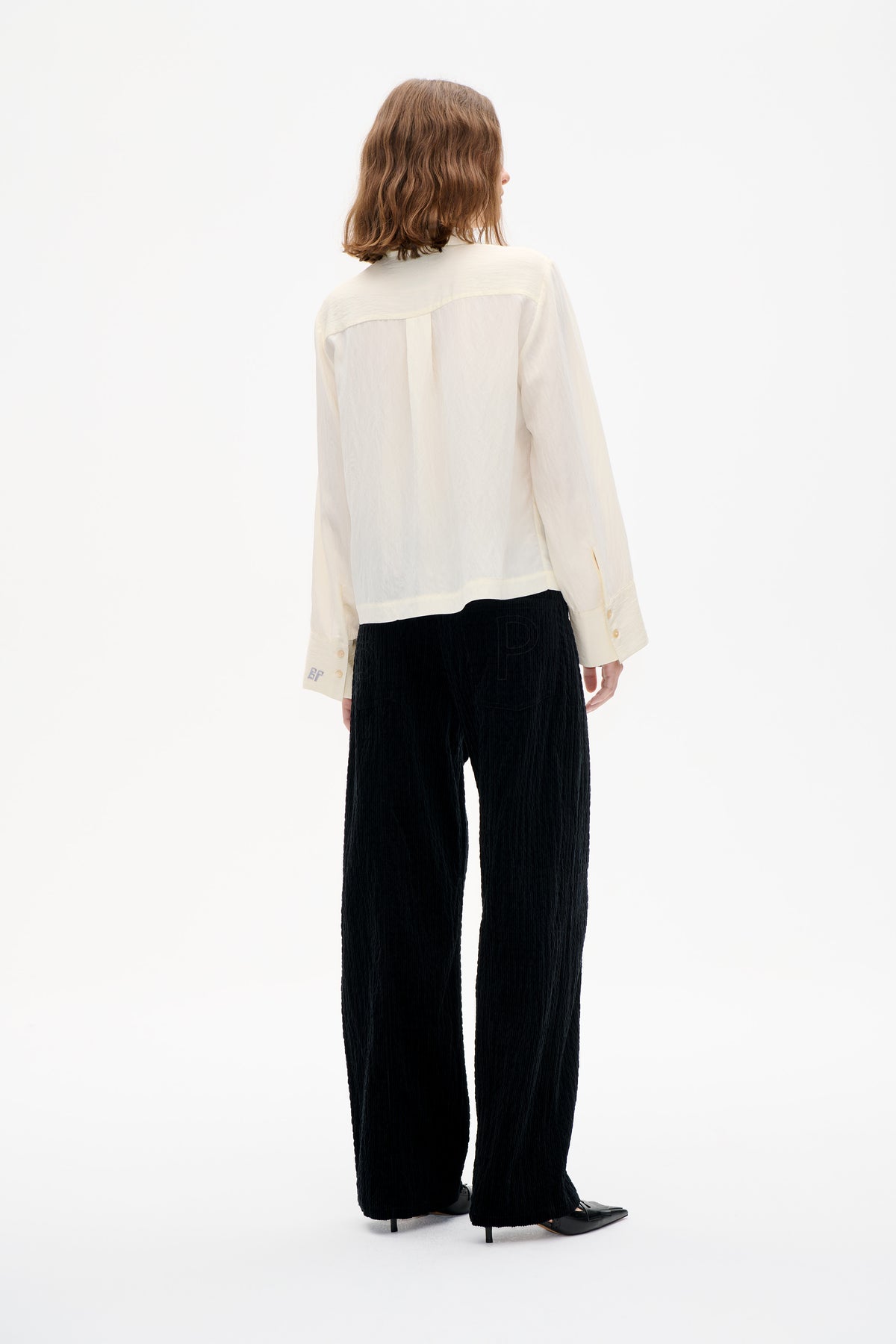 Rear view of a model wearing an ivory long-sleeved shirt with a sheer back panel and black textured wide-leg trousers.