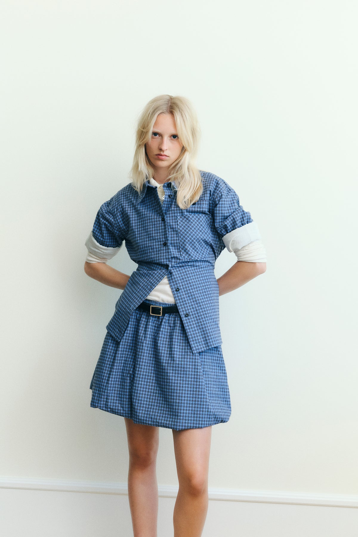 Model wearing a blue checked button-up shirt over a white top with a matching checked mini skirt against a pale wall.
