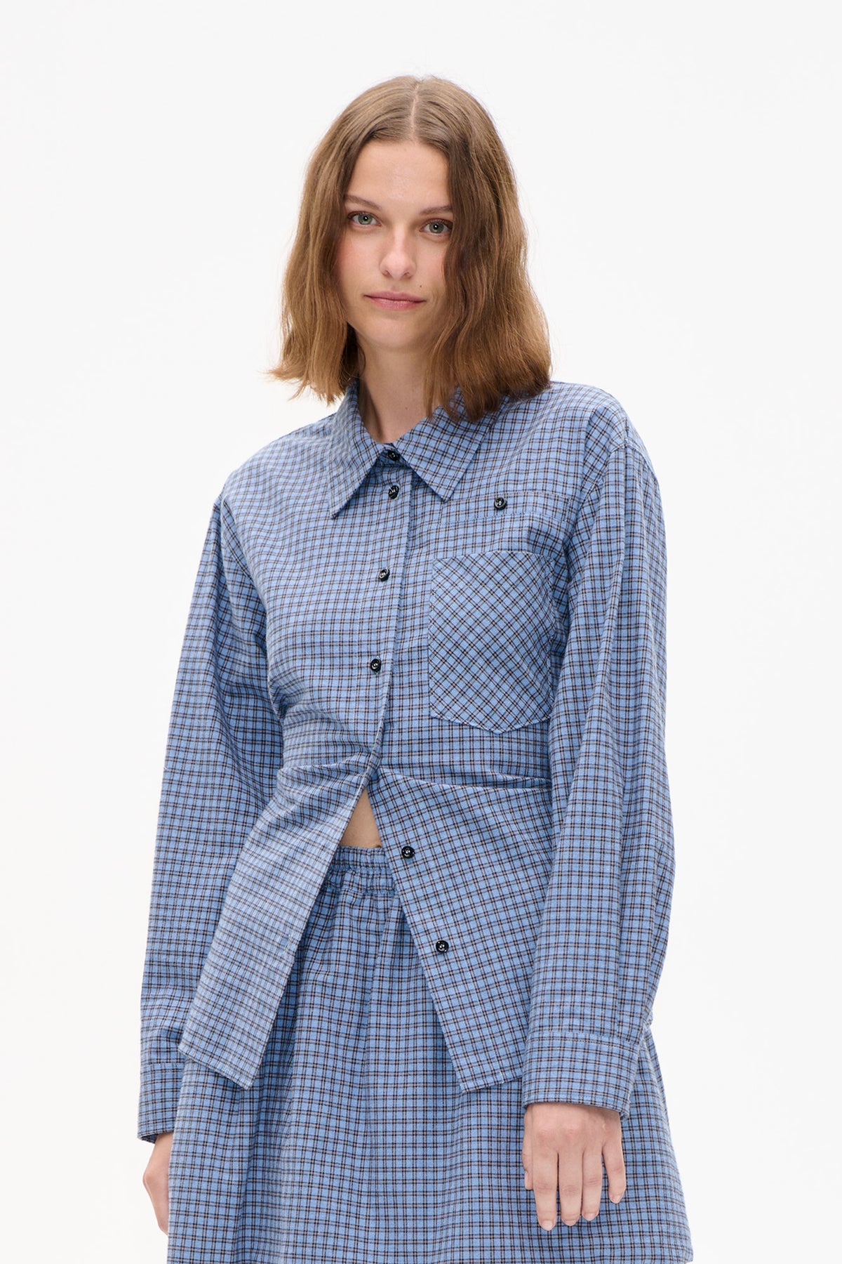 Front detail of a model wearing a light blue checked button-up shirt with a pointed collar and large diagonal-check chest pocket.