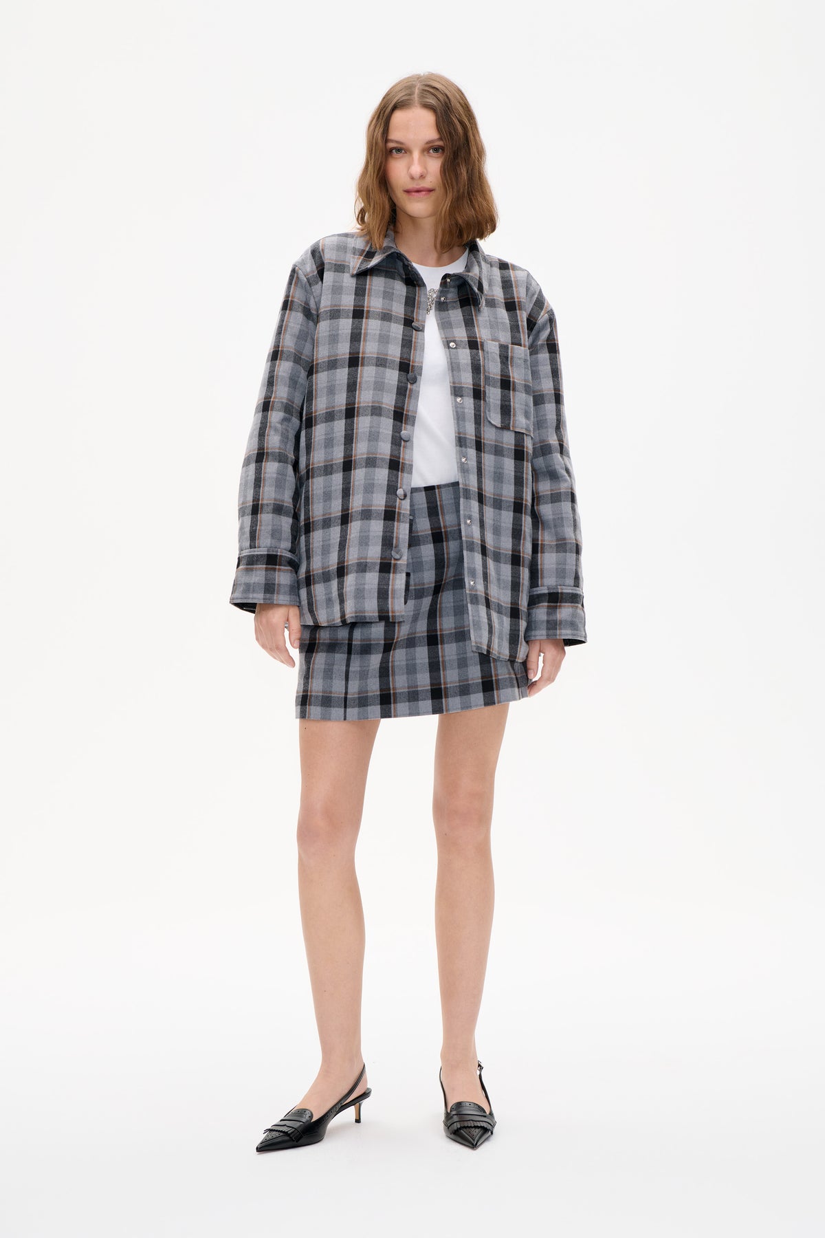 Vue de face d'un mannequin portant une surchemise à carreaux gris, noir et rouille, boutonnée devant, assortie à une mini-jupe et un top blanc.