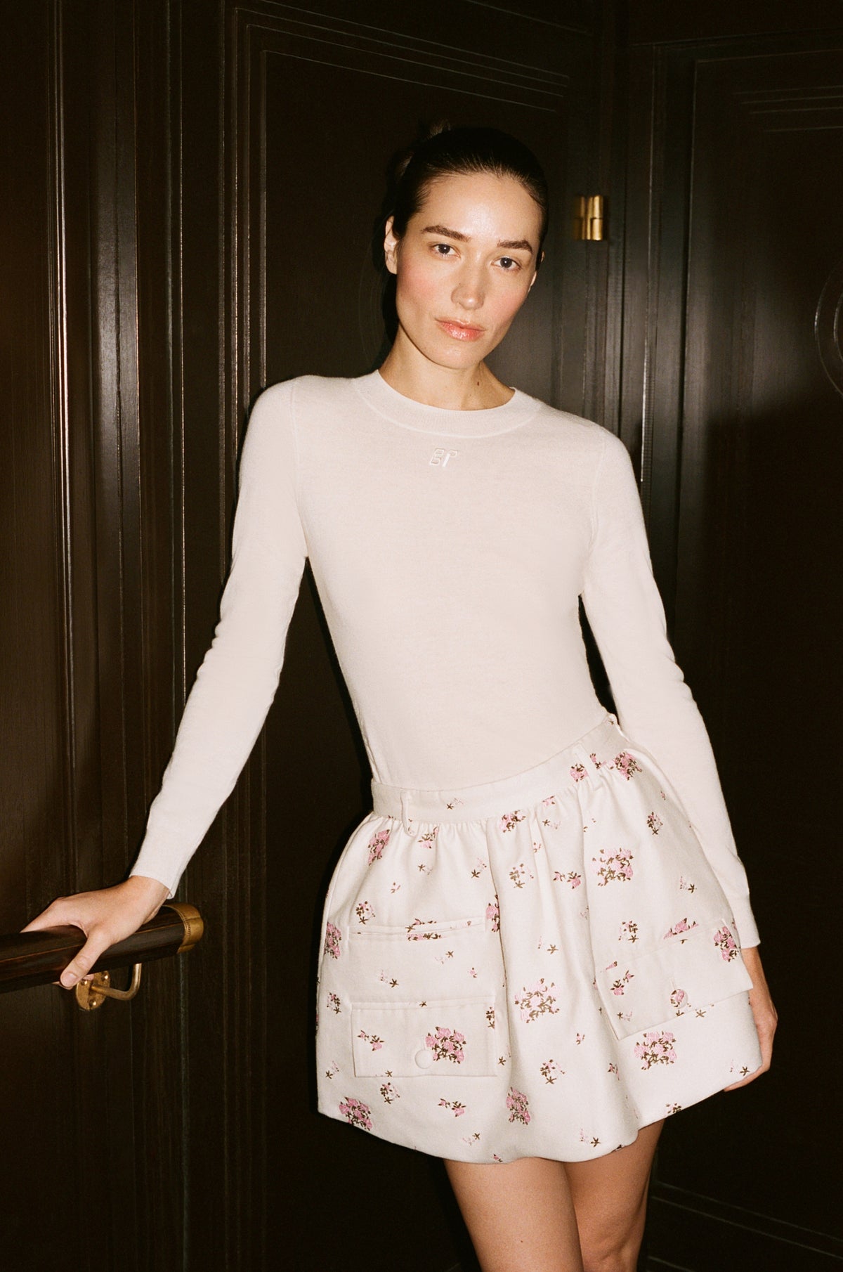 Model in an ivory long-sleeve top and pale floral shorts with large front pockets, posed against a dark paneled door.