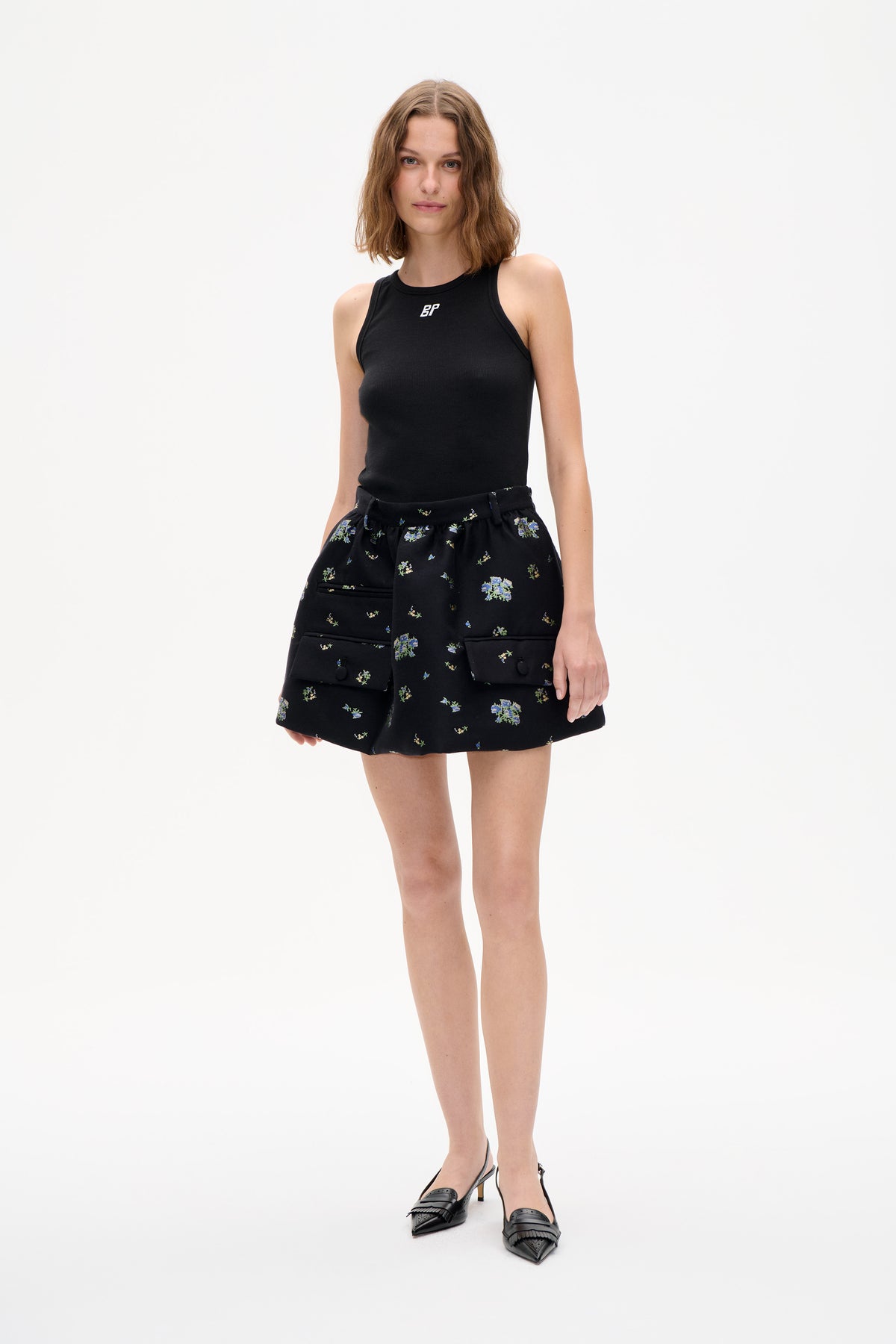 Model wearing a black sleeveless ribbed top with a white BP logo and a black floral mini skirt with large flap pockets.