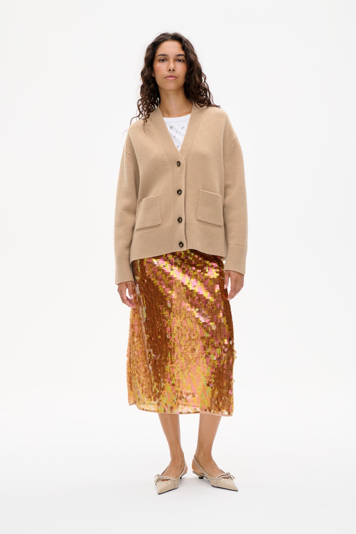 Front view of a model wearing a beige button-front knit cardigan with patch pockets over a white top and a multicoloured sequin skirt.
