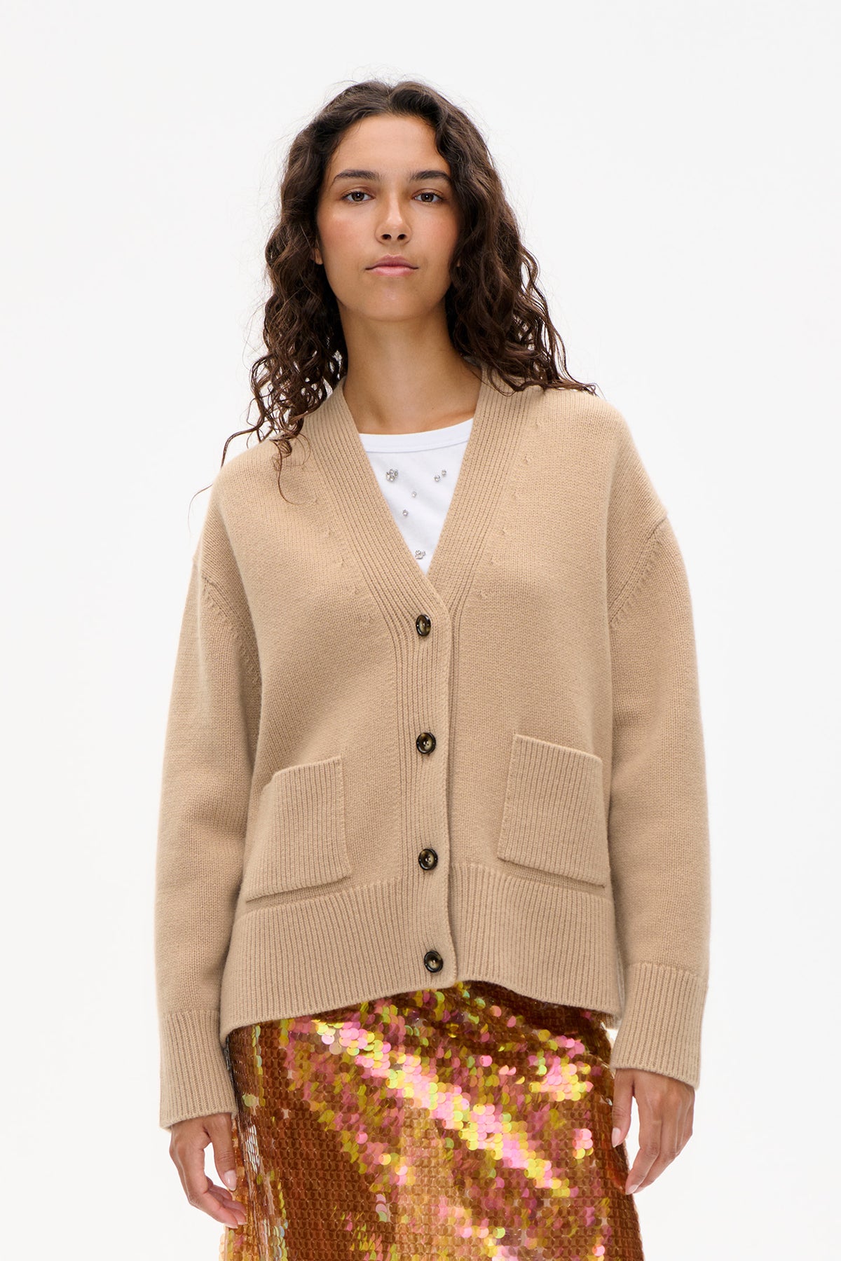 Front view of a beige knit cardigan with ribbed trim, dark buttons and patch pockets, layered over a white embellished top and multicolour sequin skirt.