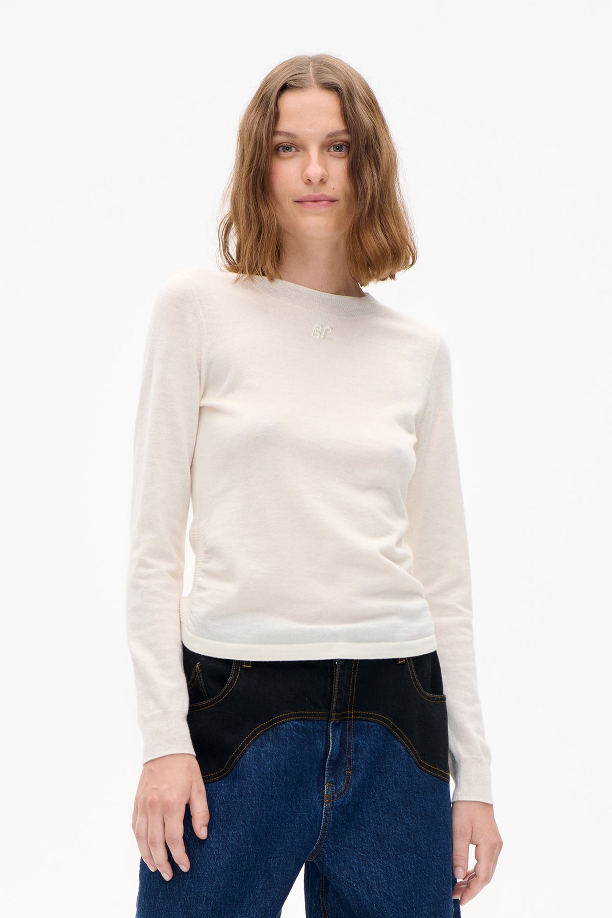 Front view of a model wearing an ivory long-sleeve knit sweater with a small tonal chest logo and dark blue jeans