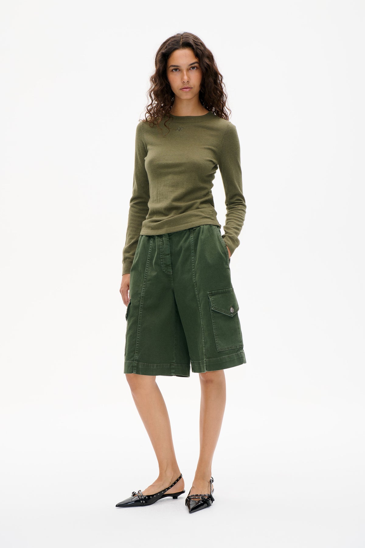 Front view of a model wearing an olive long-sleeve top, dark green knee-length cargo shorts and black pointed flats.