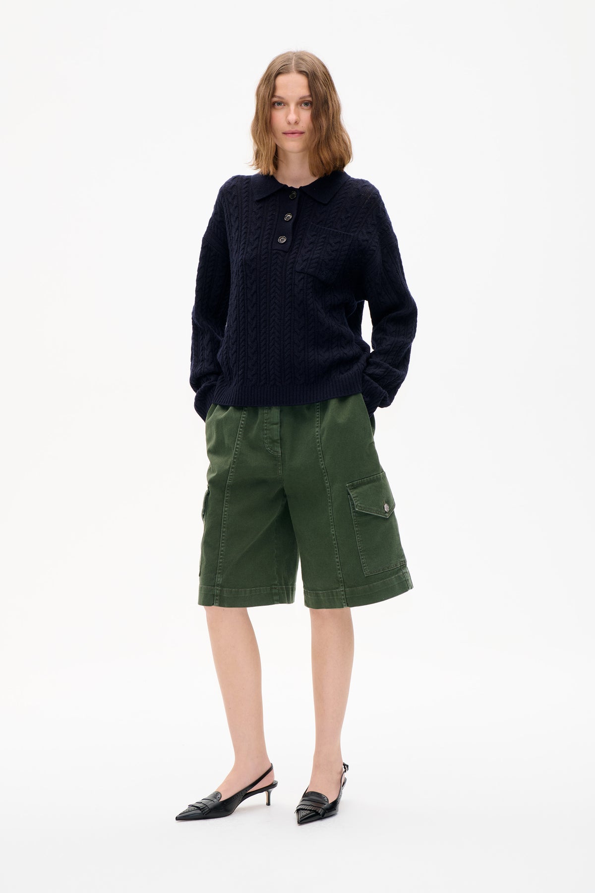 Front view of a model wearing a navy cable-knit collared sweater, green cargo shorts and black slingback heels against a white background.