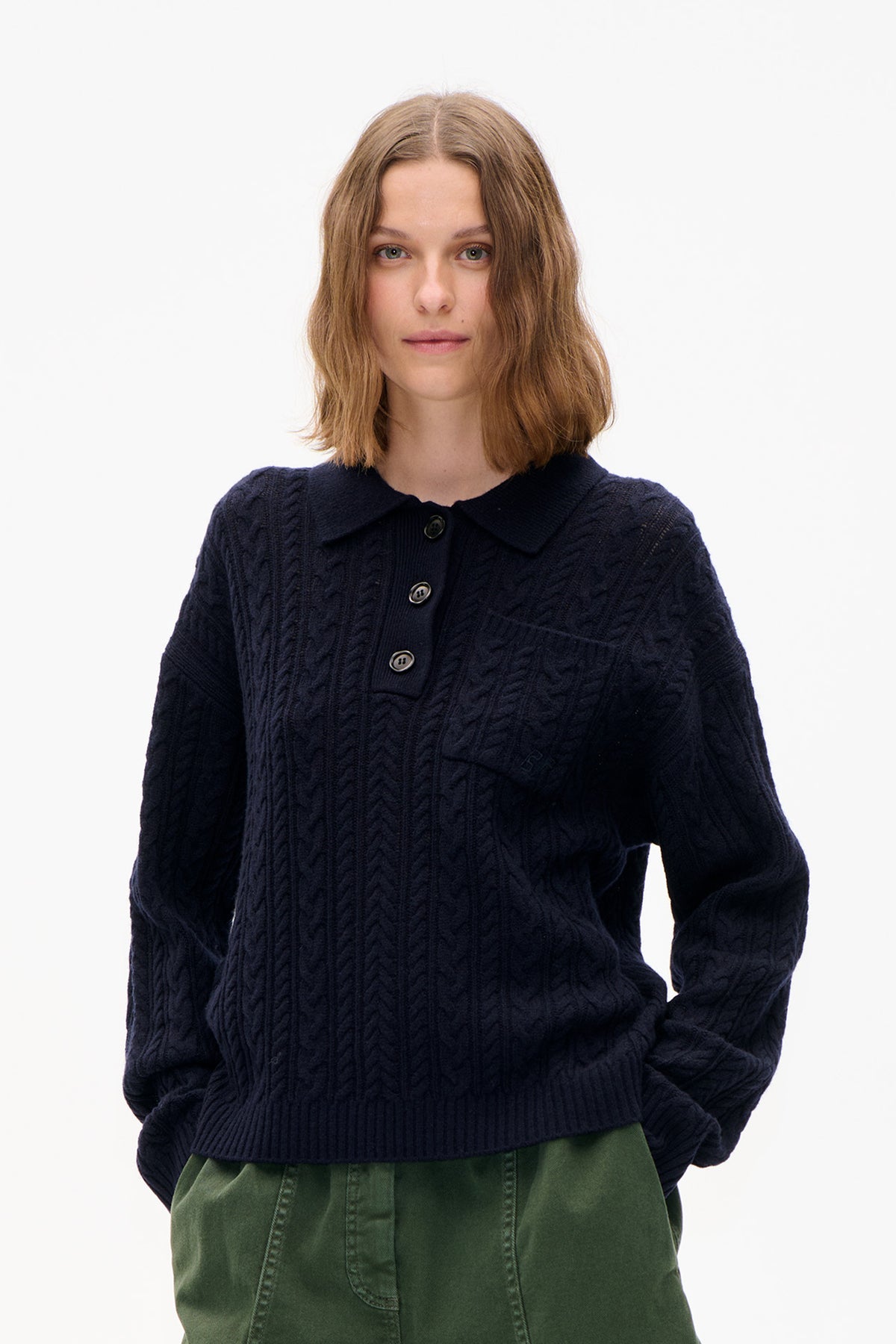 Front view of a model wearing a navy cable-knit cardigan with a wide collar and buttons, paired with green trousers.