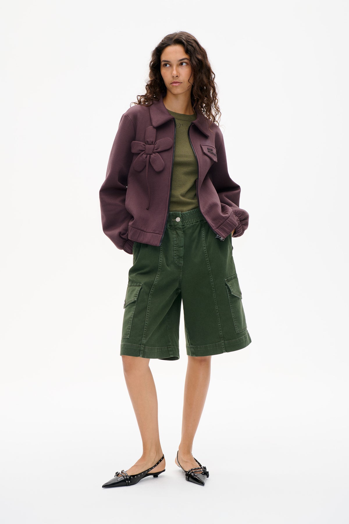 Model wearing a plum zip-front jacket with an oversized bow detail, an olive top and green cargo shorts against a white backdrop.