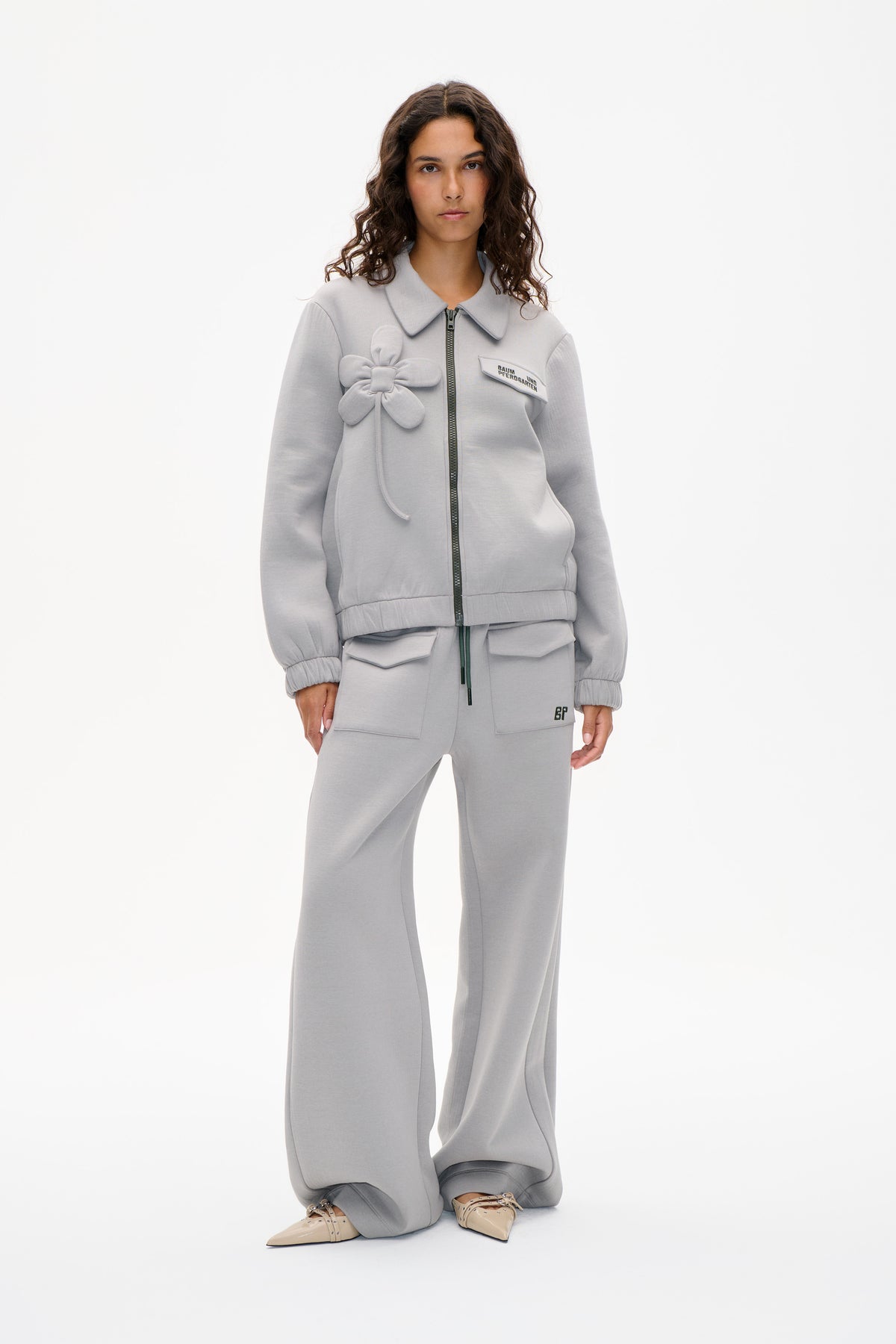 Model wearing a light grey zip-front jacket with an oversized collar and flower appliqué, paired with matching wide-leg trousers.