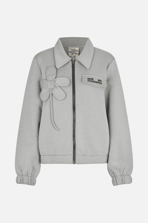 Front view of a light grey zip-up jacket with a pointed collar, gathered cuffs, a flower appliqué and a logo flap pocket.
