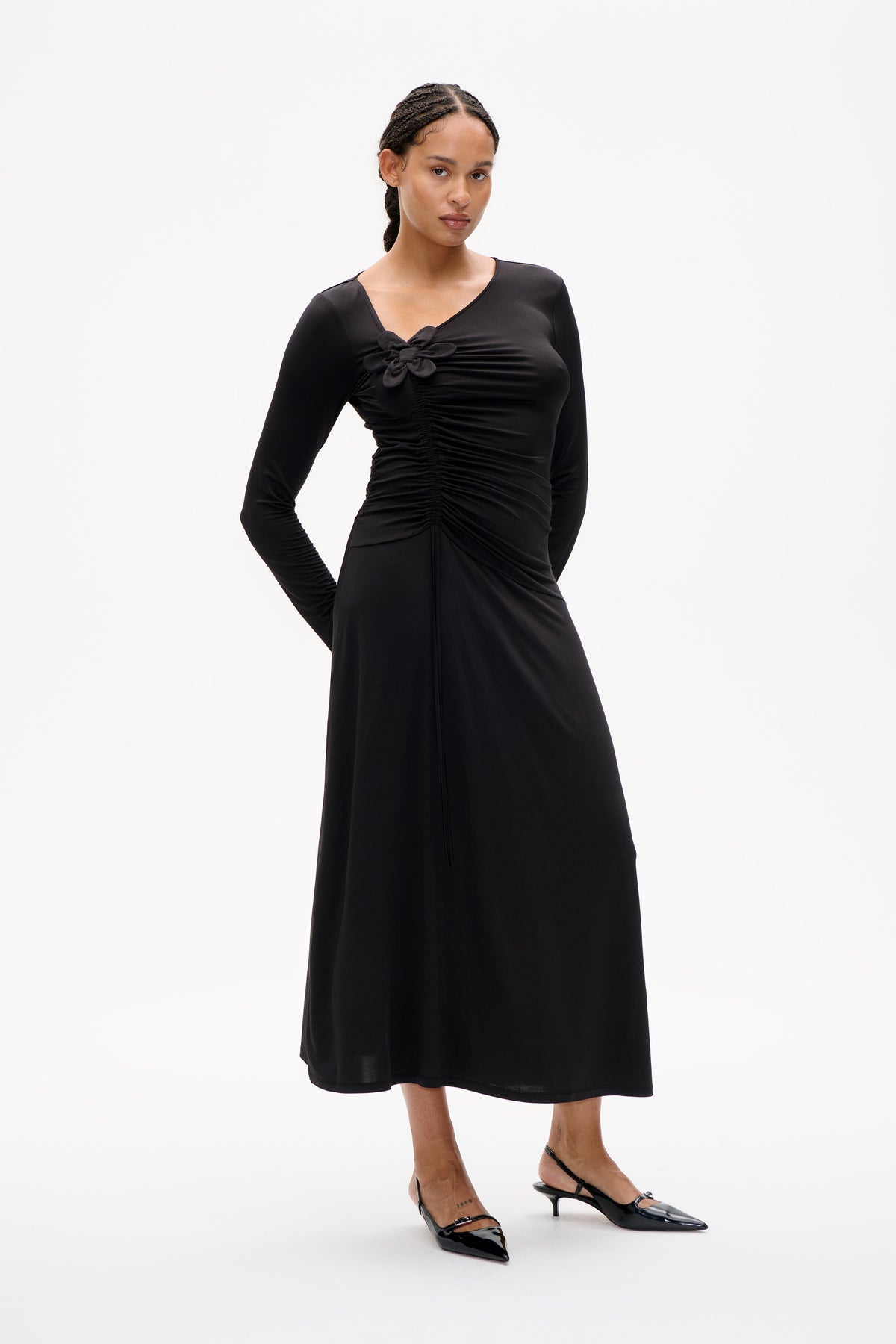 Front view of a black long-sleeve dress with a V-neck, gathered ruching and a fabric flower detail at the chest.