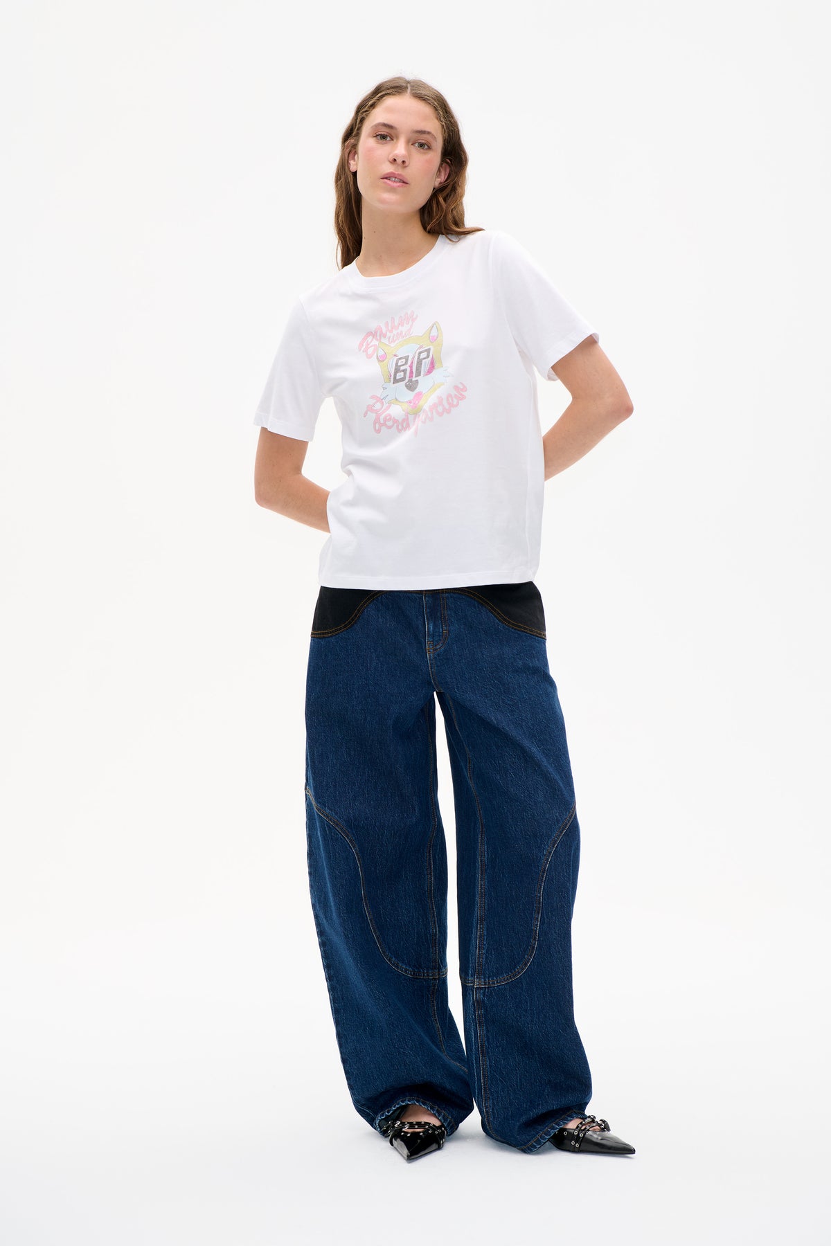 Model draagt een wit T-shirt met korte mouwen, een pastelgekleurde cartoonprint en Baum und Pferdgarten-belettering, gecombineerd met donkerblauwe jeans met wijde pijpen.