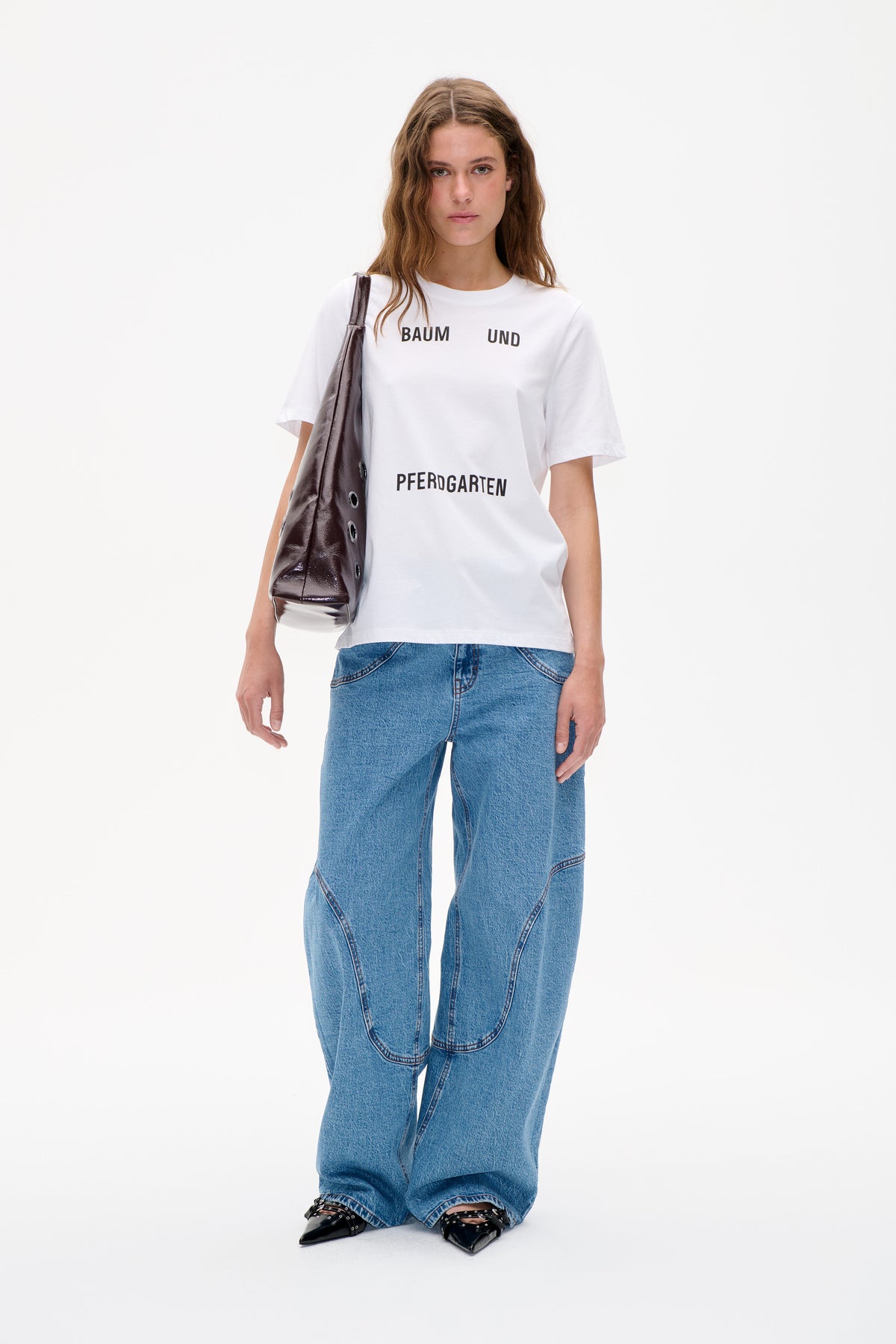 Model wearing a white T-shirt with black “BAUM UND PFERDGARTEN” lettering, blue wide-leg jeans and a glossy dark shoulder bag.