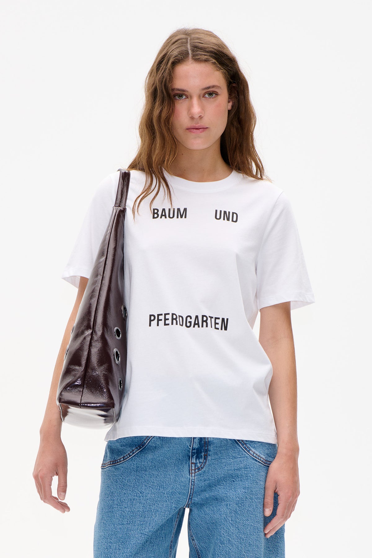 Model wearing a white short-sleeved T-shirt with bold black Baum und Pferdgarten lettering, blue jeans and a glossy dark shoulder bag.