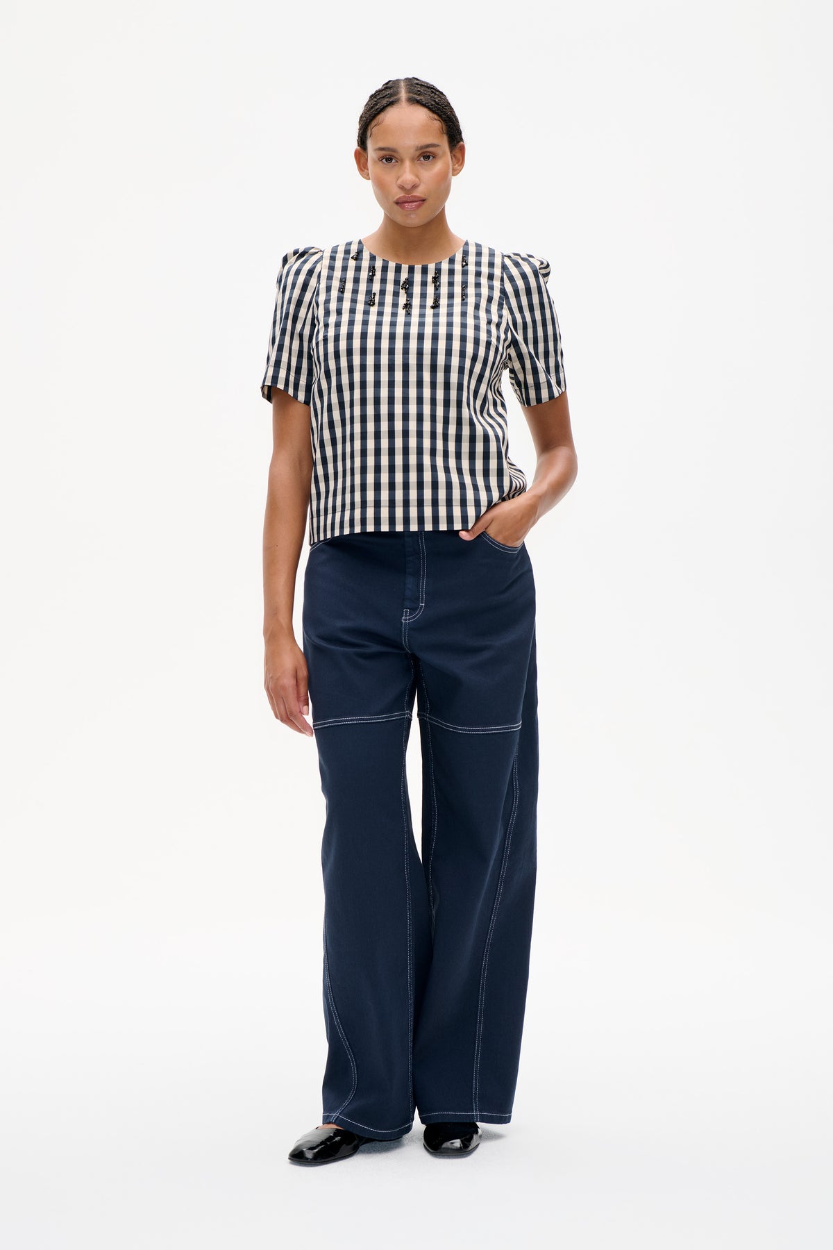 Model trägt ein kariertes Top in Marine und Creme mit gepufften Schultern, kurzen Ärmeln und schwarzen Perlenverzierungen, kombiniert mit weiten marineblauen Hosen.