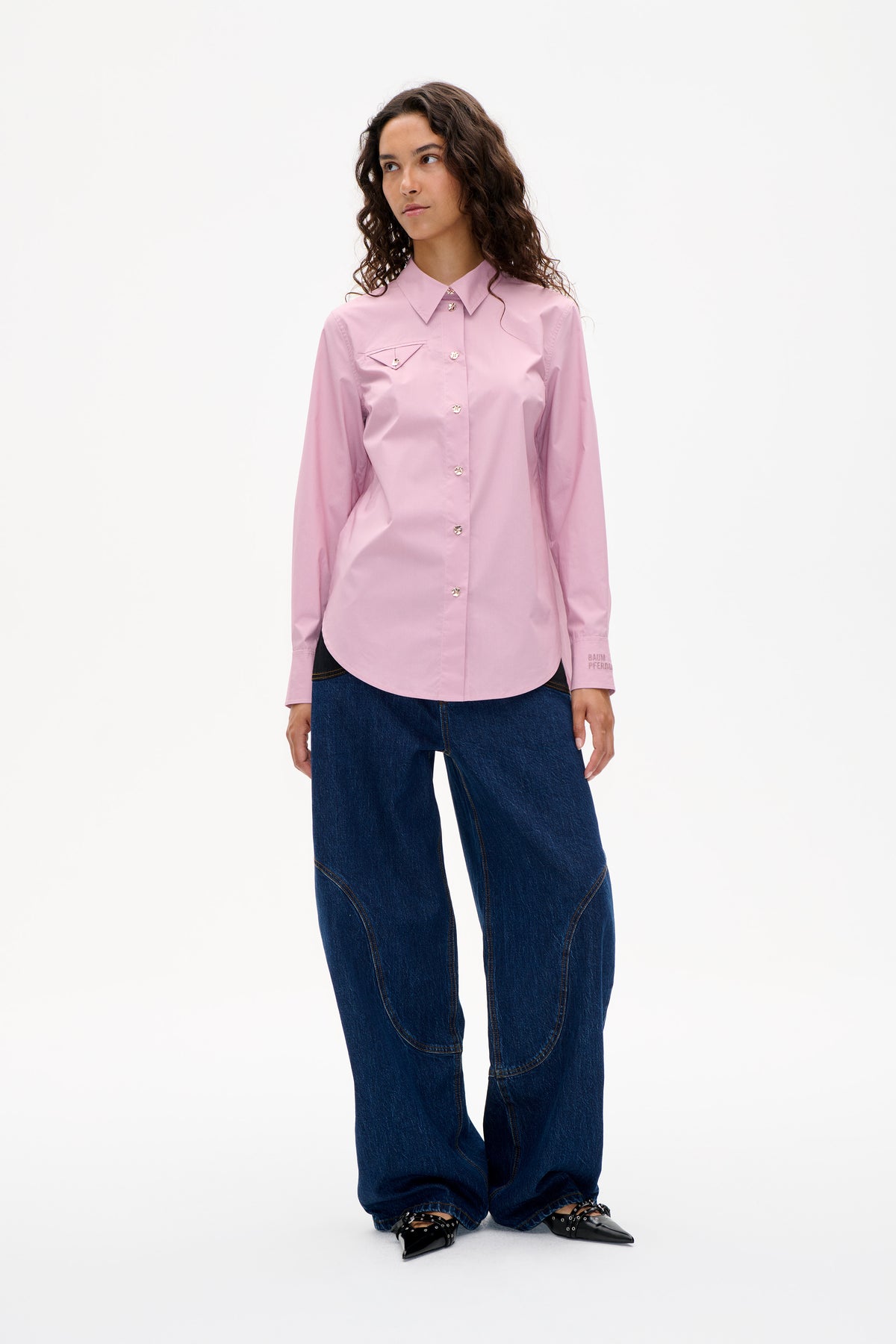 Vorderansicht eines Models, das eine zartrosa Bluse mit spitzem Kragen und aufgesetzter Brusttasche trägt, kombiniert mit dunklen, weit geschnittenen Jeans.