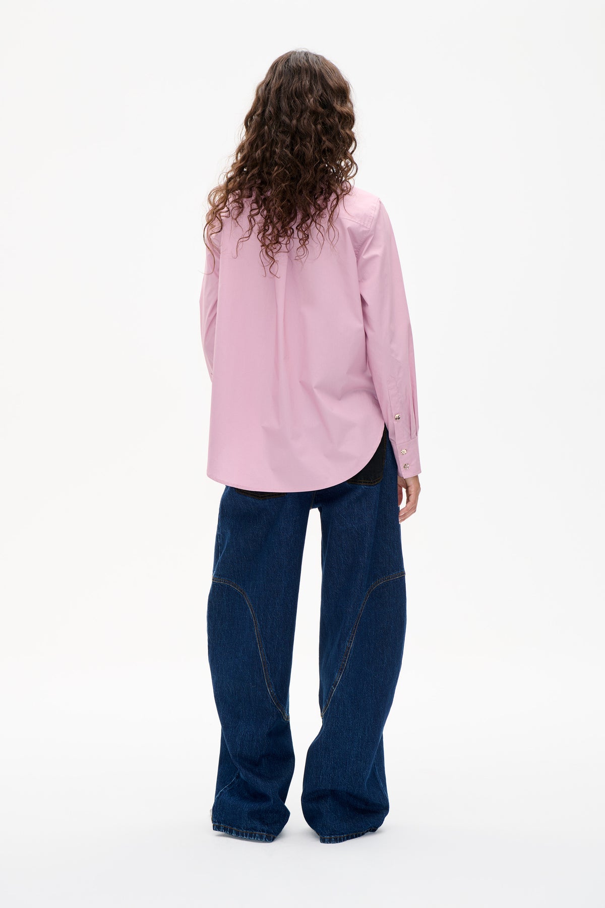 Rückansicht eines Models, das ein schlichtes hellrosa Langarmshirt mit geknöpften Manschetten und dunkelblaue Jeans mit weitem Bein trägt.