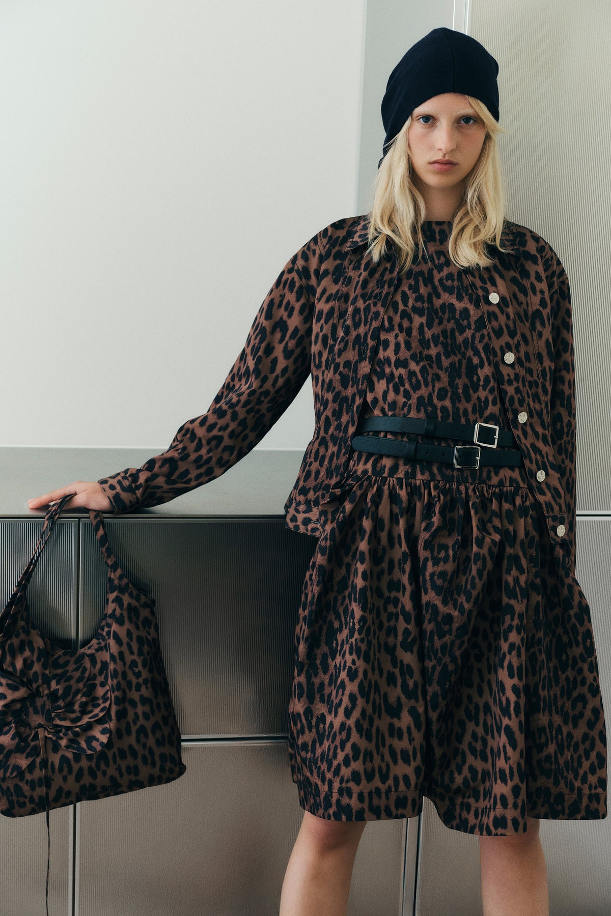 Campaign portrait of a model in a brown-and-black leopard-print button-front dress with a black double belt and beanie; another model is partly visible at left.
