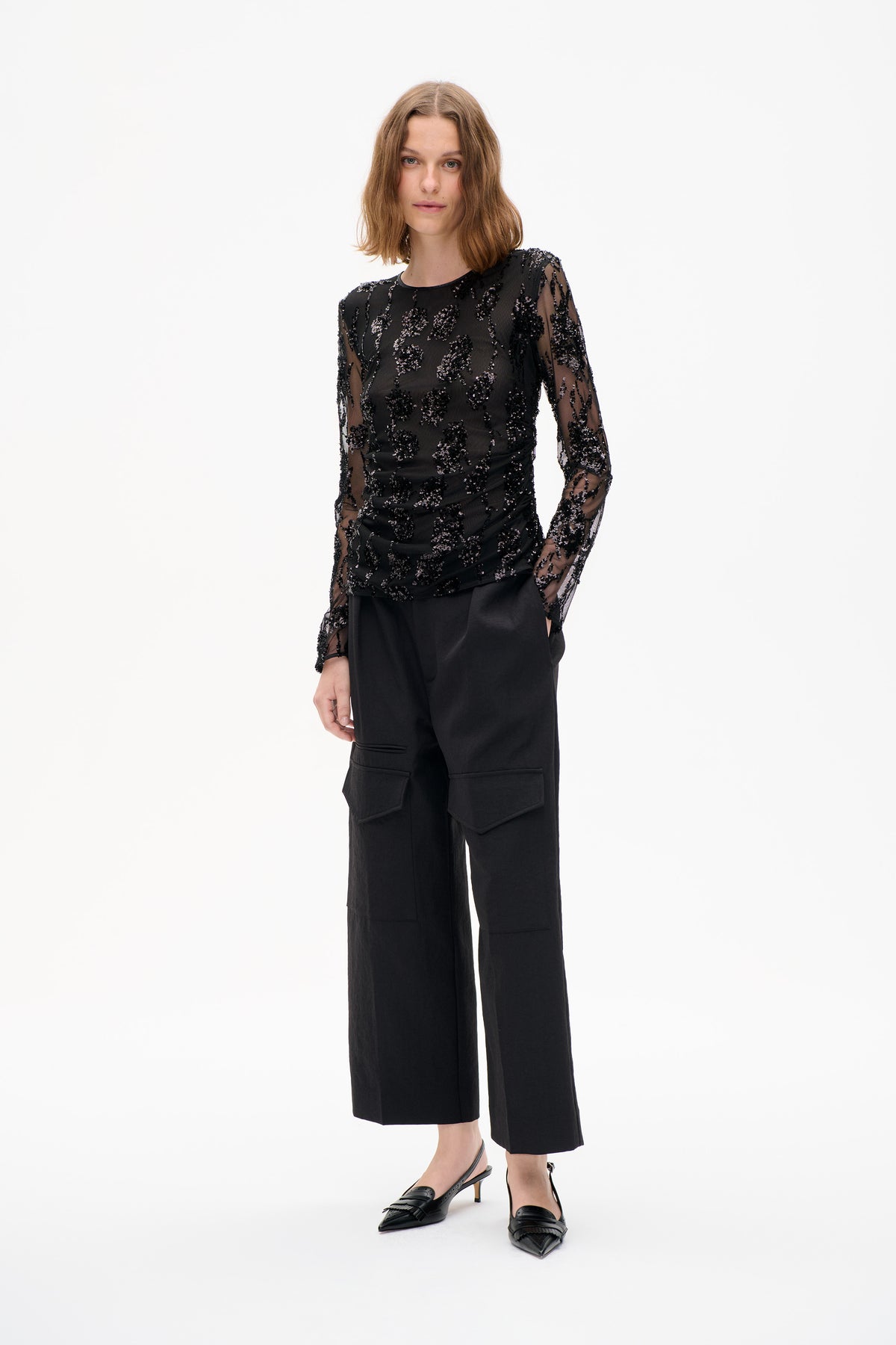 Model wearing a sheer black long-sleeved top with sequined motifs and black wide-leg cargo trousers, front view against a white background.