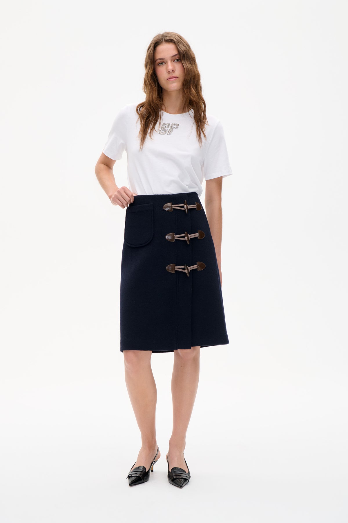 Model trägt ein weißes T‑Shirt mit kurzen Ärmeln und funkelndem Brustmonogramm sowie einen marineblauen Rock mit Knebelverschluss vorn und aufgesetzter Tasche.