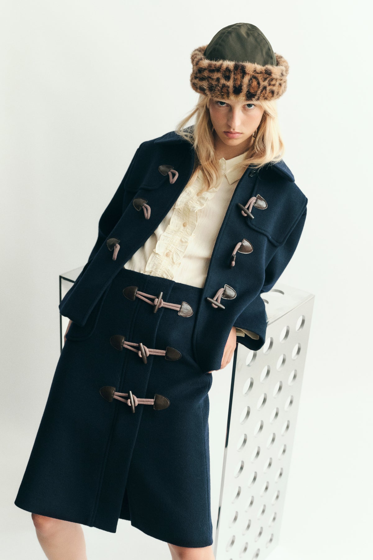 Kampagnenporträt eines Models in einem marineblauen Dufflecoat mit pinken Kordelverschlüssen, einer cremefarbenen Rüschenbluse und einem braunen, gemusterten Pelzhut.