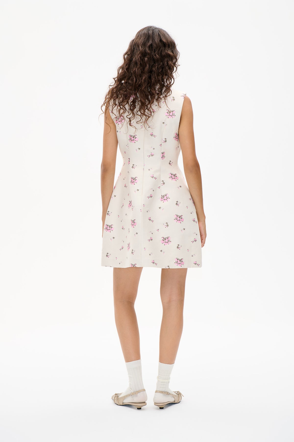 Rückansicht eines ärmellosen cremefarbenen Minikleids mit verstreutem rosa-braunem Blumenmuster, gestylt mit weißen Socken und beigen Ballerinas.