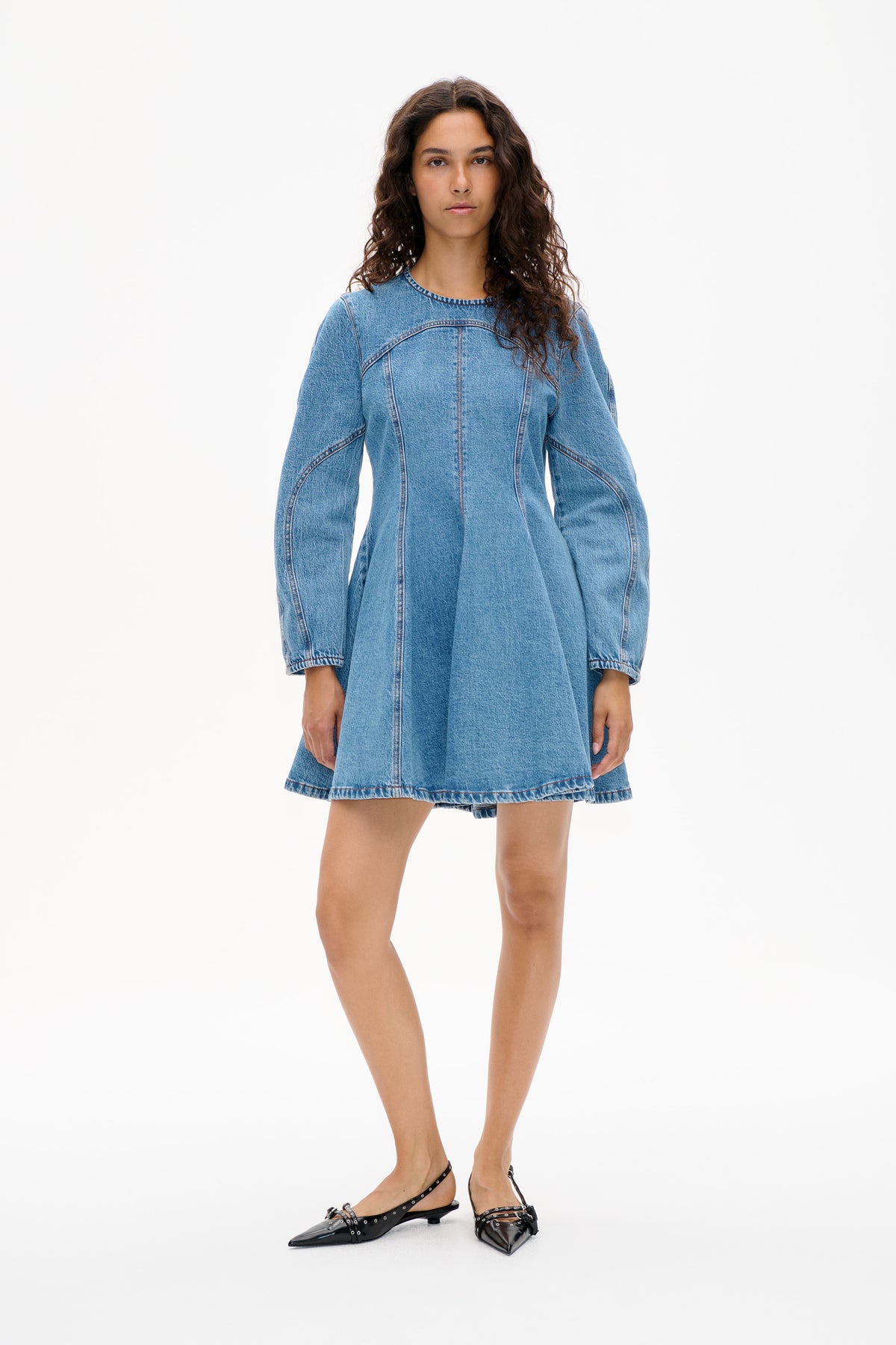 Model wearing a light blue denim mini dress with long sleeves, a curved front yoke and panel seams, shown from the front.