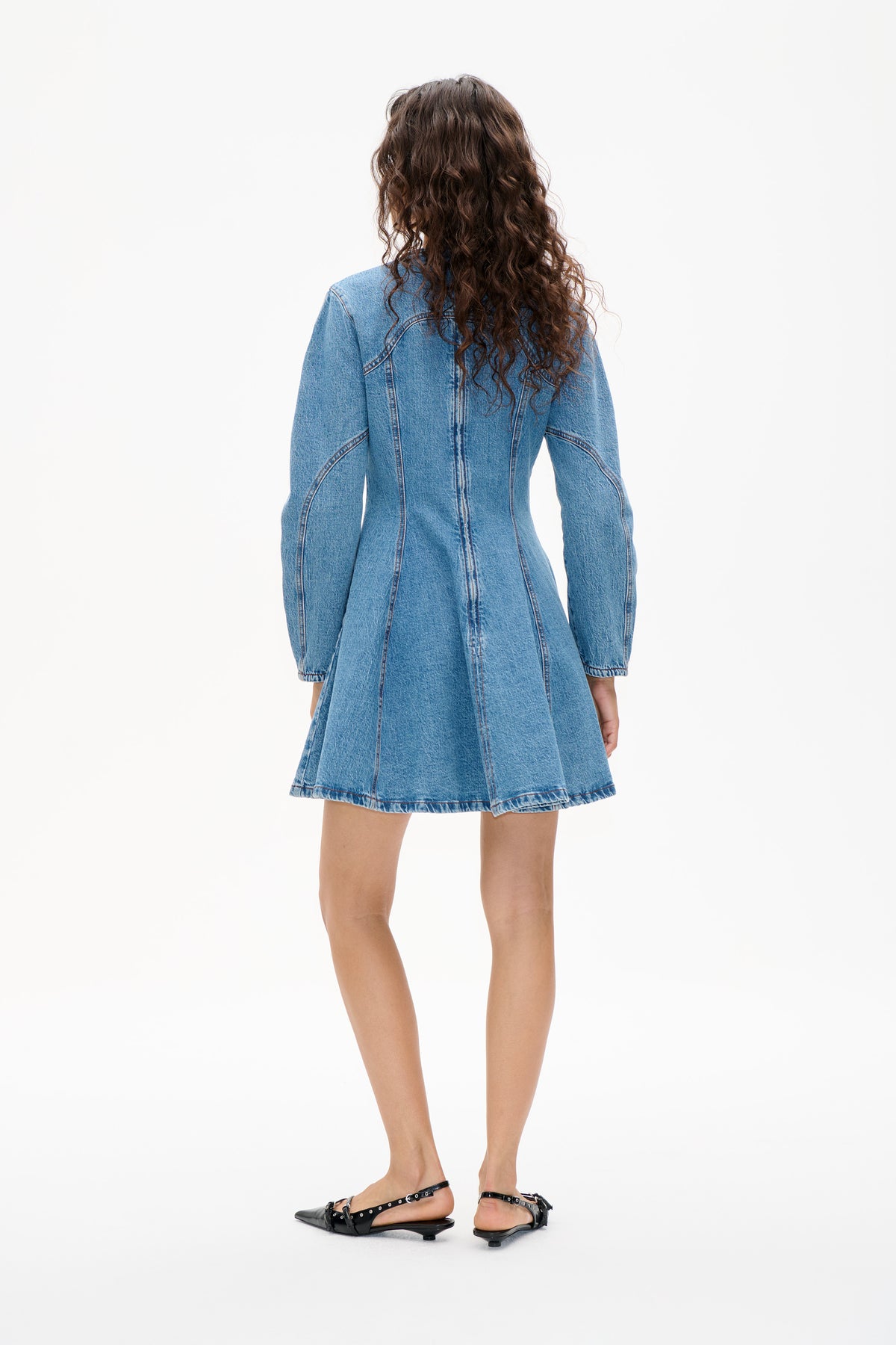 Achteraanzicht van een model dat een lichtblauwe denimjurk met lange mouwen draagt, met een uitlopende rok en gebogen contrastnaden.