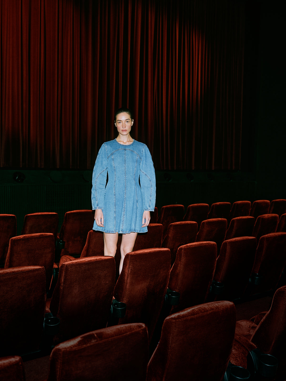 Model tussen bordeauxrode theaterstoelen in een lichtblauwe denim mini-jurk met lange mouwen en gebogen paneelnaden.