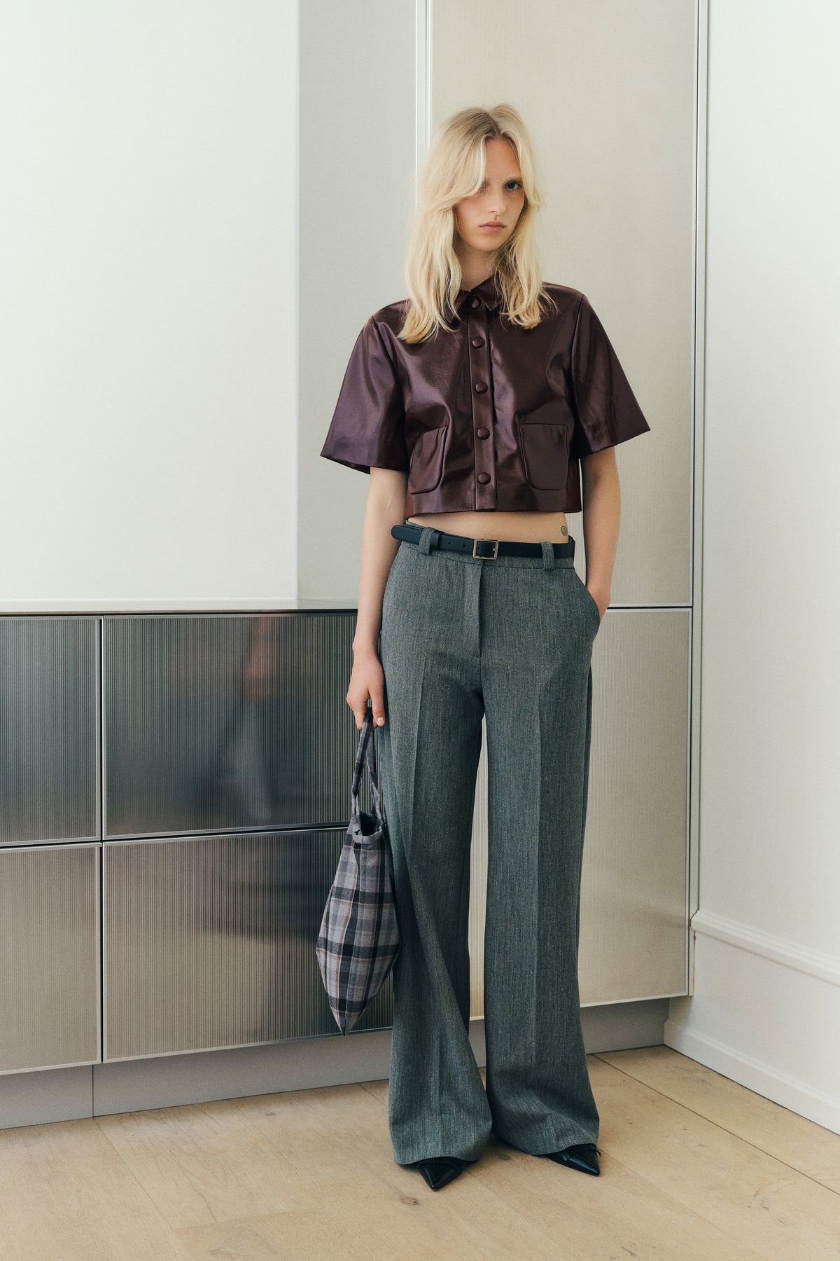 Kampagnenporträt eines Models, das eine glänzende, pflaumenfarbene kurzärmlige Hemdbluse mit durchgehender Knopfleiste, eine graue, weit geschnittene Hose trägt und einen karierten Shopper hält.