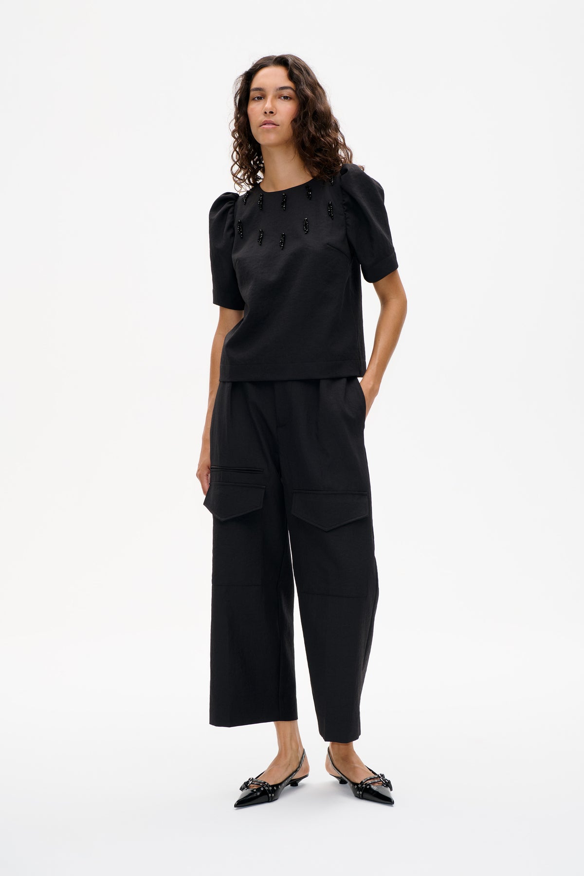 Model trägt ein schwarzes Top mit Puffärmeln und Perlenverzierung vorne, kombiniert mit einer kurz geschnittenen, weiten Hose mit großen aufgesetzten Taschen.