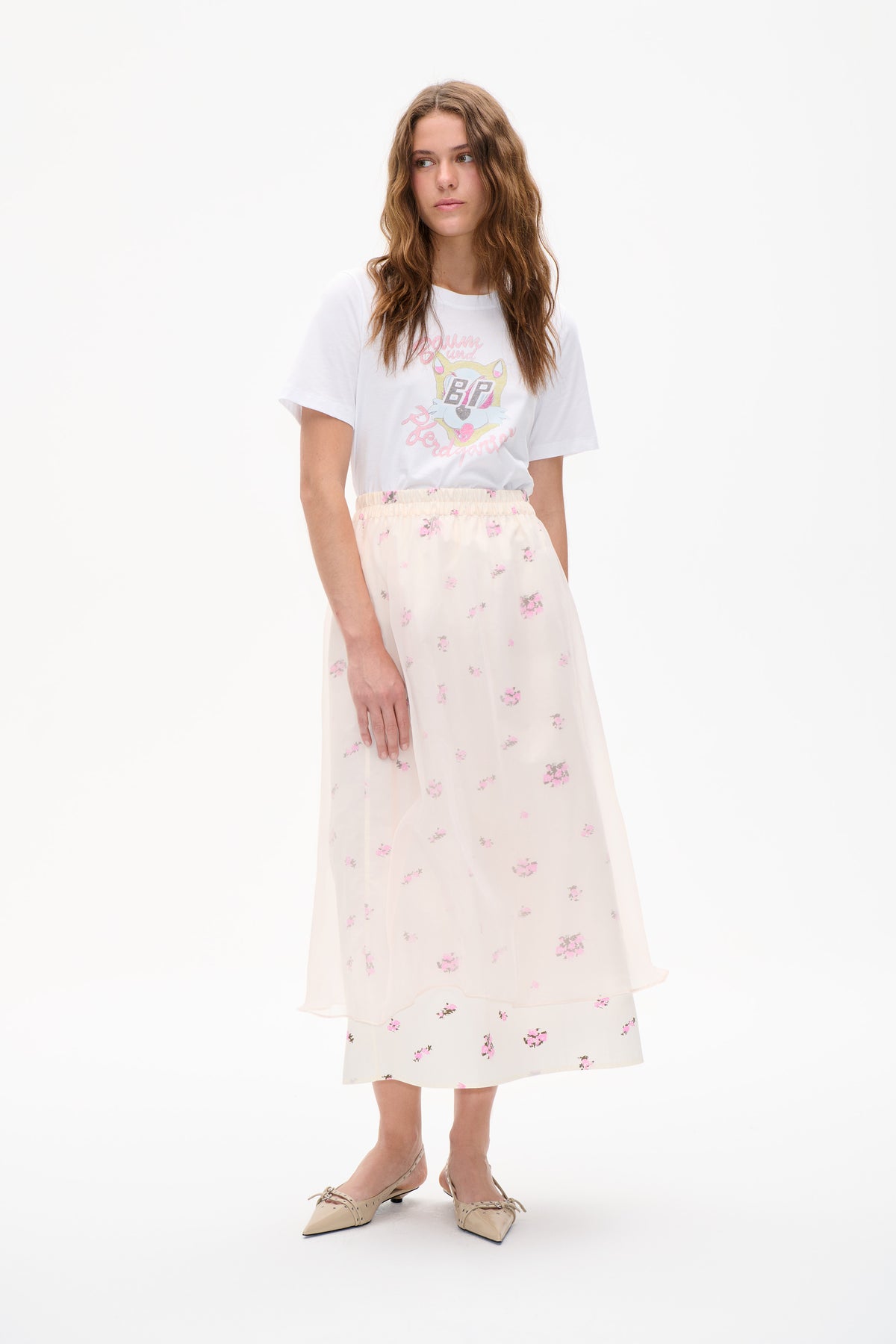 Model trägt ein weißes, bedrucktes T-Shirt mit buntem Katzenemblem und einen transparenten, elfenbeinfarbenen Rock mit Blumenmuster vor weißem Studiohintergrund.