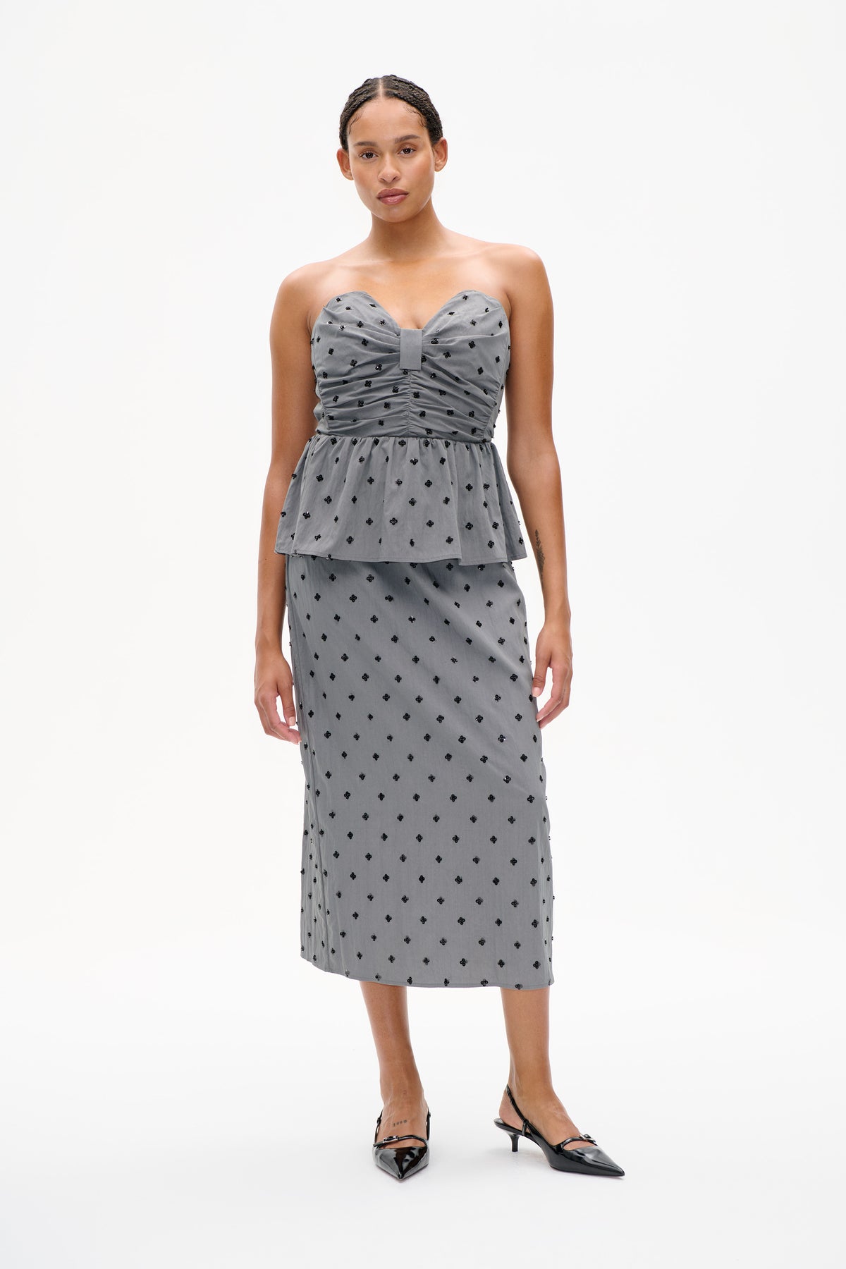 Front view of a model wearing a grey strapless midi dress with a ruched sweetheart bodice, peplum overlay and black floral embellishments.