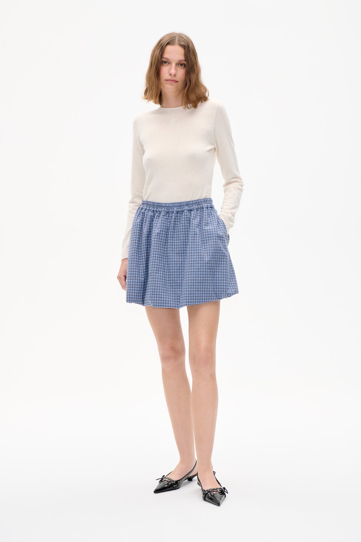 Model trägt einen elfenbeinfarbenen Rundhalspullover mit kleinem Brustmonogramm, einen gerafften blau-karierten Minirock und schwarze, spitze Slipper vor weißem Hintergrund.