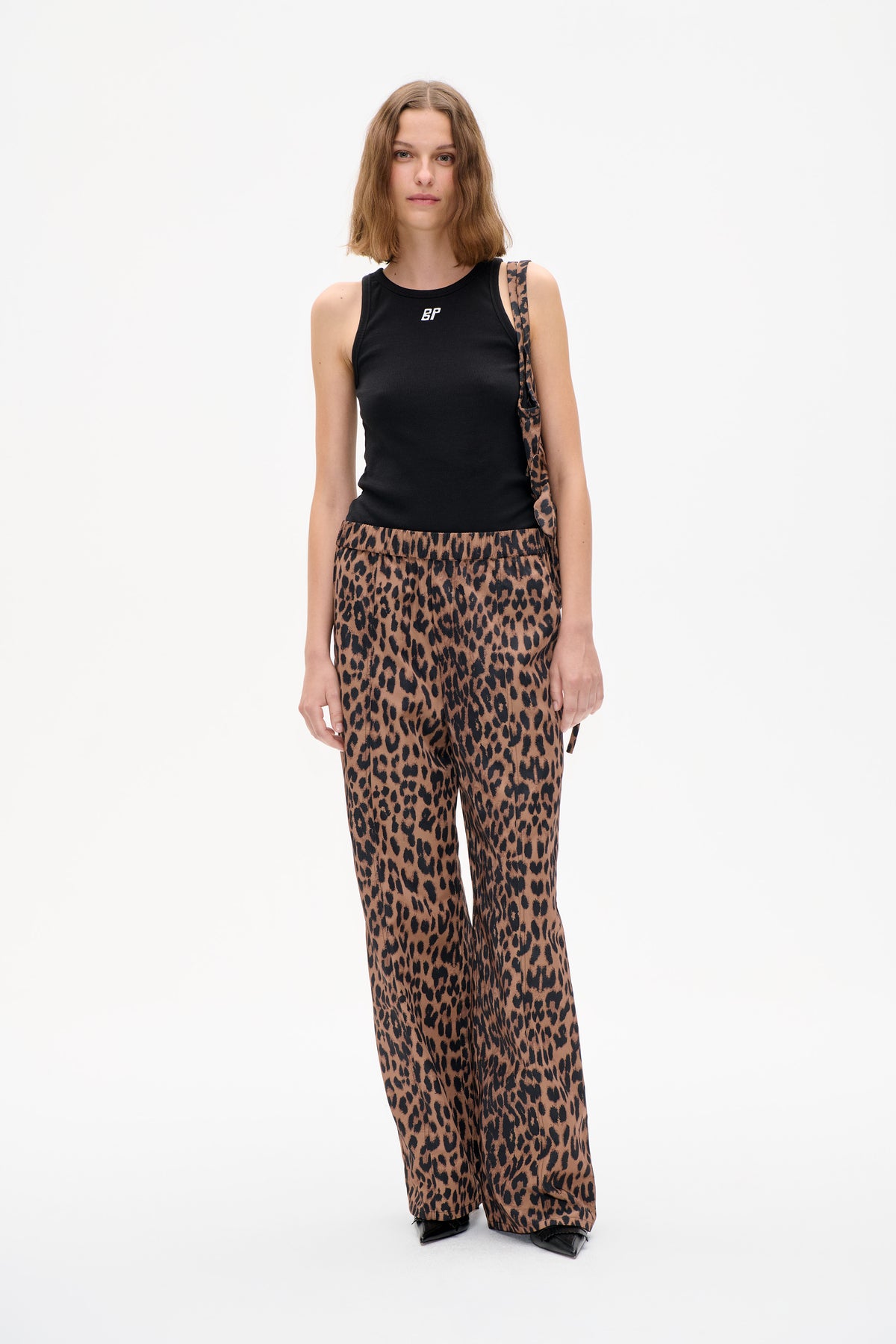 Model trägt ein schwarzes, ärmelloses Top mit kleinem weißen Logo und weite Hosen mit braun-schwarzem Leopardenmuster. Frontalansicht.