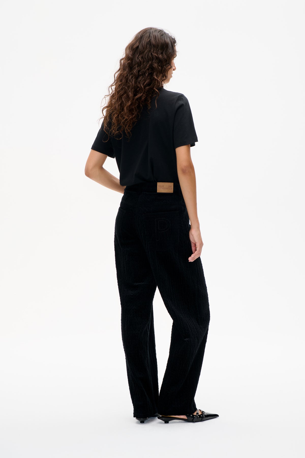 Rückansicht eines Models in schwarzem T-Shirt mit kurzen Ärmeln und schwarzer Kordhose mit beigefarbenem Bundetikett.