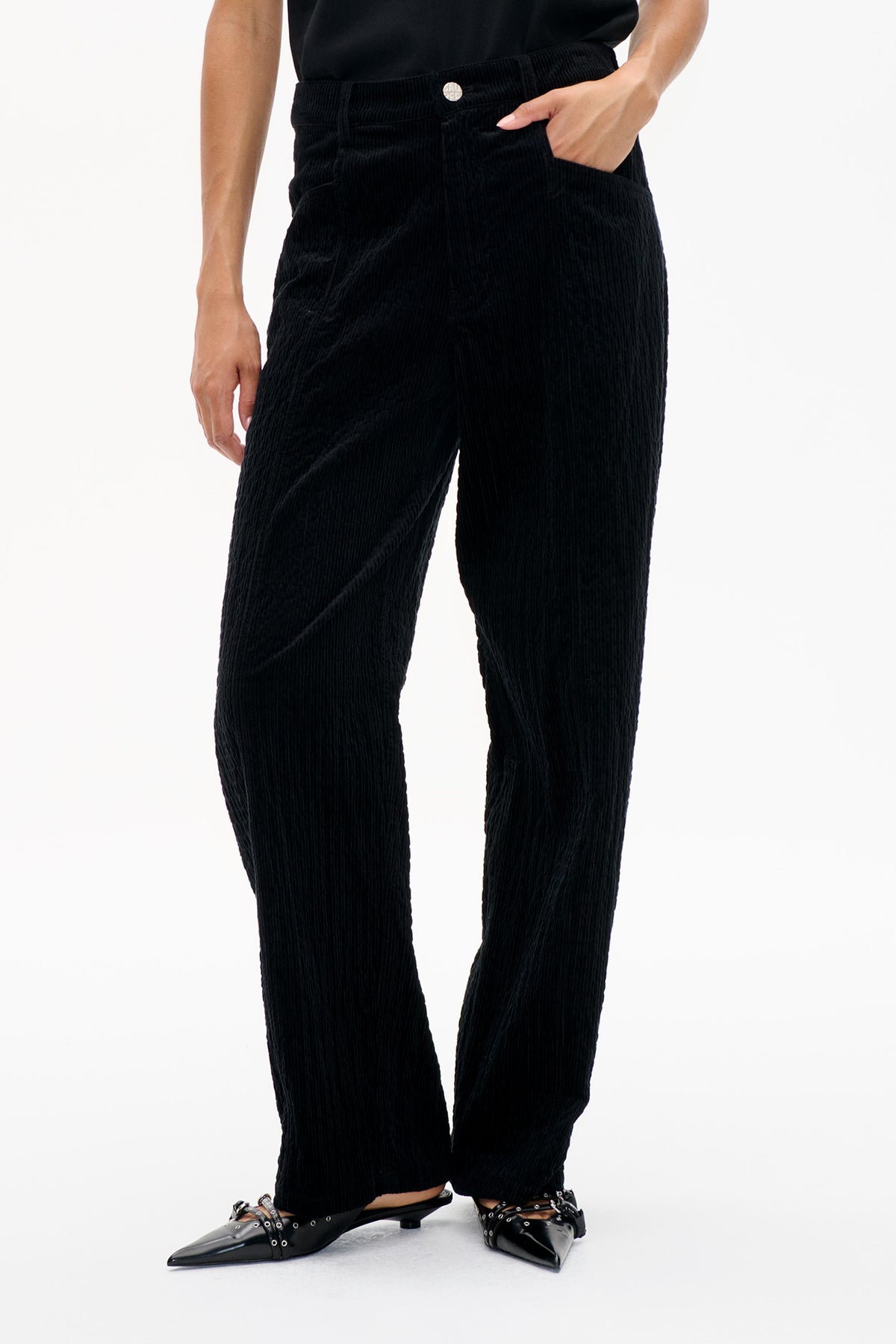 Frontdetail einer schwarzen, gerippten Cordhose mit knöpfbarem Bund und Seitentaschen, kombiniert mit spitzen schwarzen Flats.